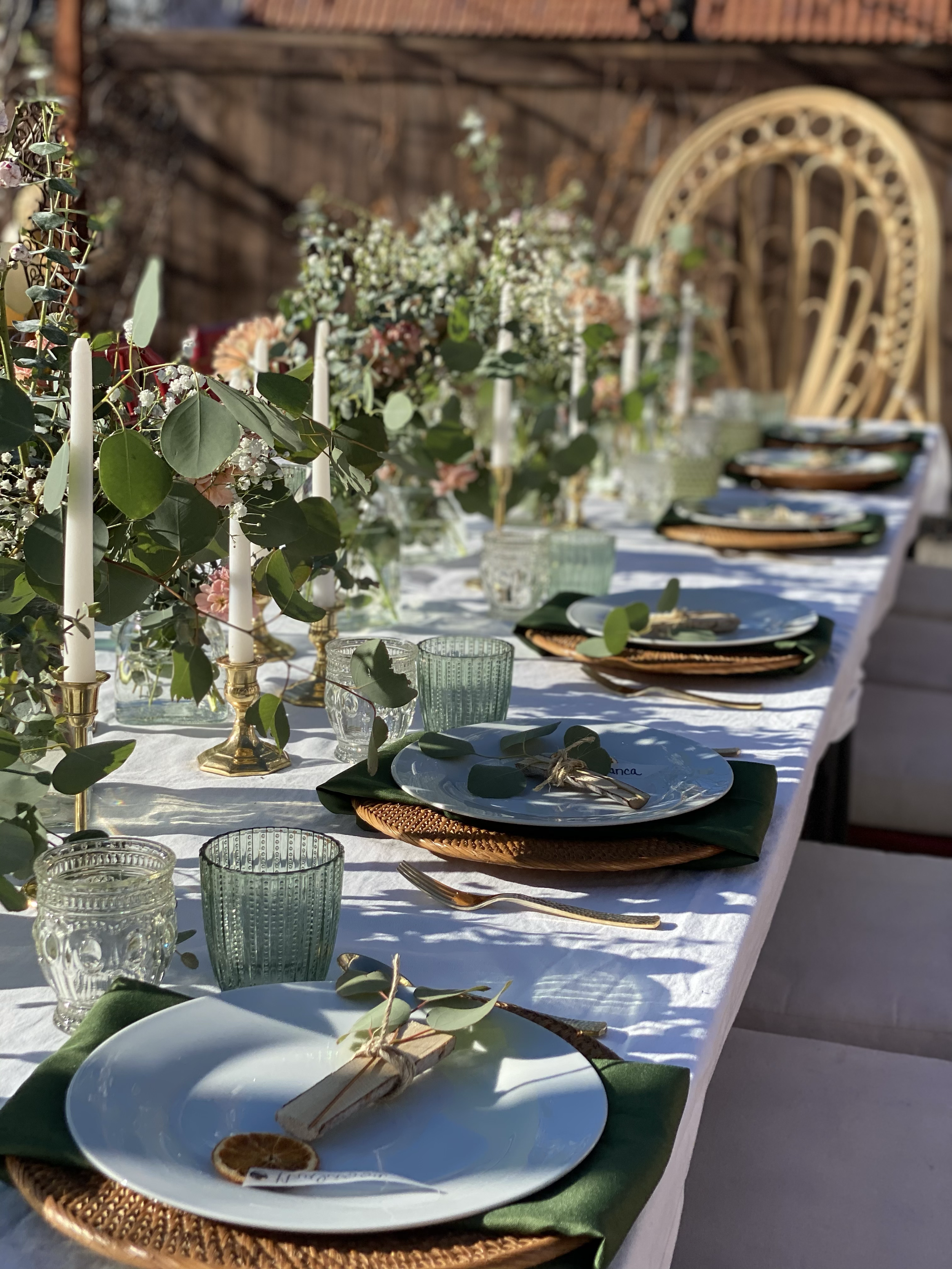 A long dining table set outdoors with elegant tableware, glassware, and floral centerpieces, surrounded by a rustic wooden background.
