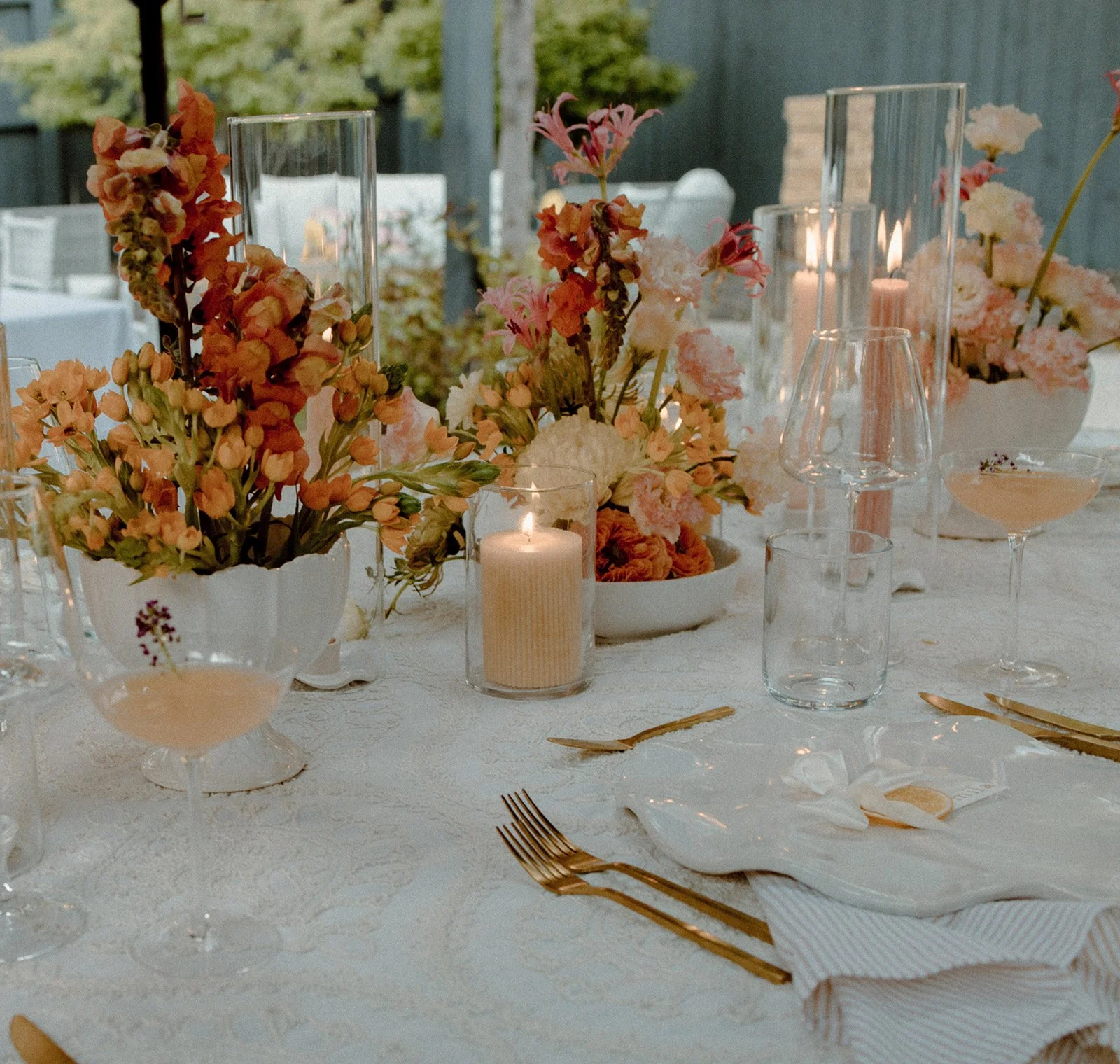 Elegant table setting with floral arrangements, candles, glassware, and gold utensils on a white tablecloth, outdoor setting with greenery in the background.
