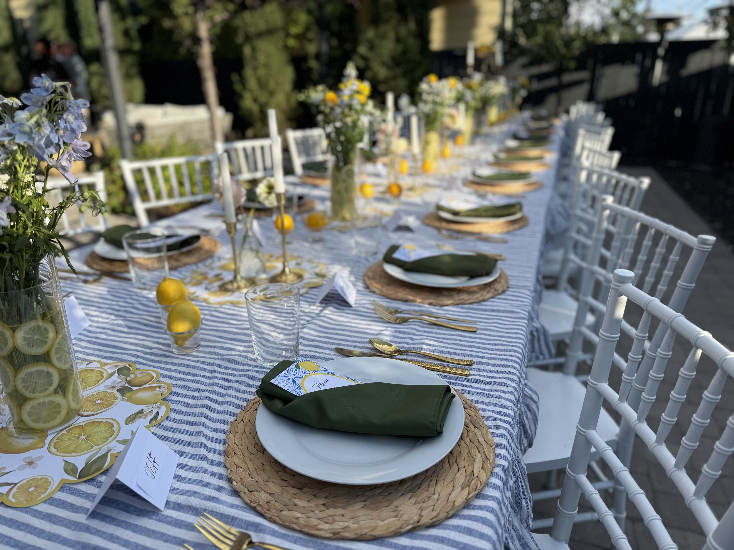 A long outdoor dining table set for a meal with white chairs, surrounded by greenery, decorated with floral centerpieces, lemons, candles, and place settings with green napkins and menus on a blue and white striped tablecloth.
