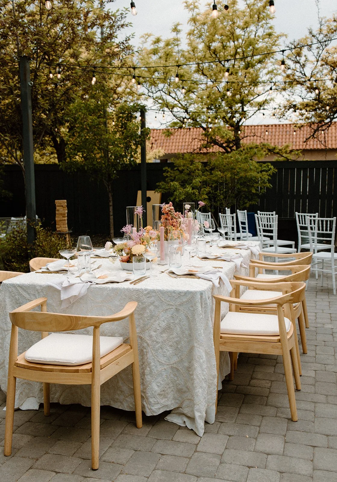 An outdoor dining table decorated with pink, white, and peach flowers in glass vases, surrounded by wooden chairs with white cushions. String lights hang above, and trees and a brick building are in the background.