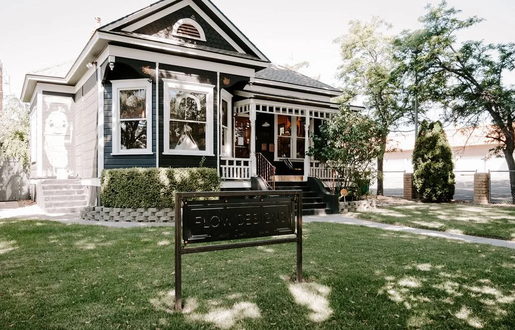 Front view of a charming, dark-colored house with white trim, a small porch with stairs, and a lawn with trees and shrubbery.