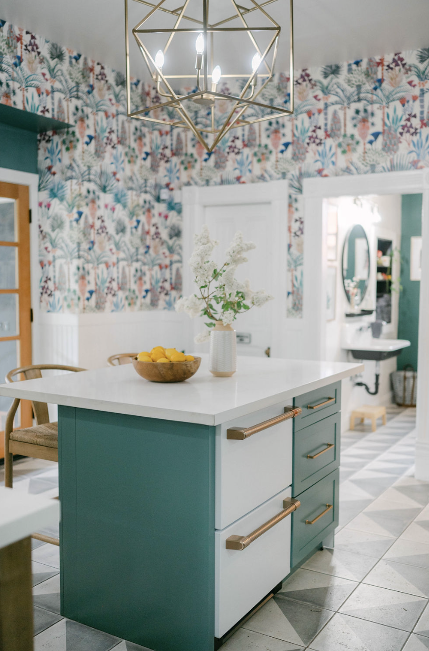 A modern kitchen island with a white marble top and teal cabinetry with brass handles, decorated with a white vase of white flowers and a bowl of lemons. The background features a colorful botanical wallpaper and a geometric gold chandelier hanging f