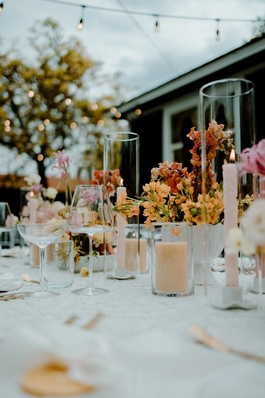 Outdoor event table decorated with floral centerpieces, candles, and glassware, with string lights overhead and trees in the background.
