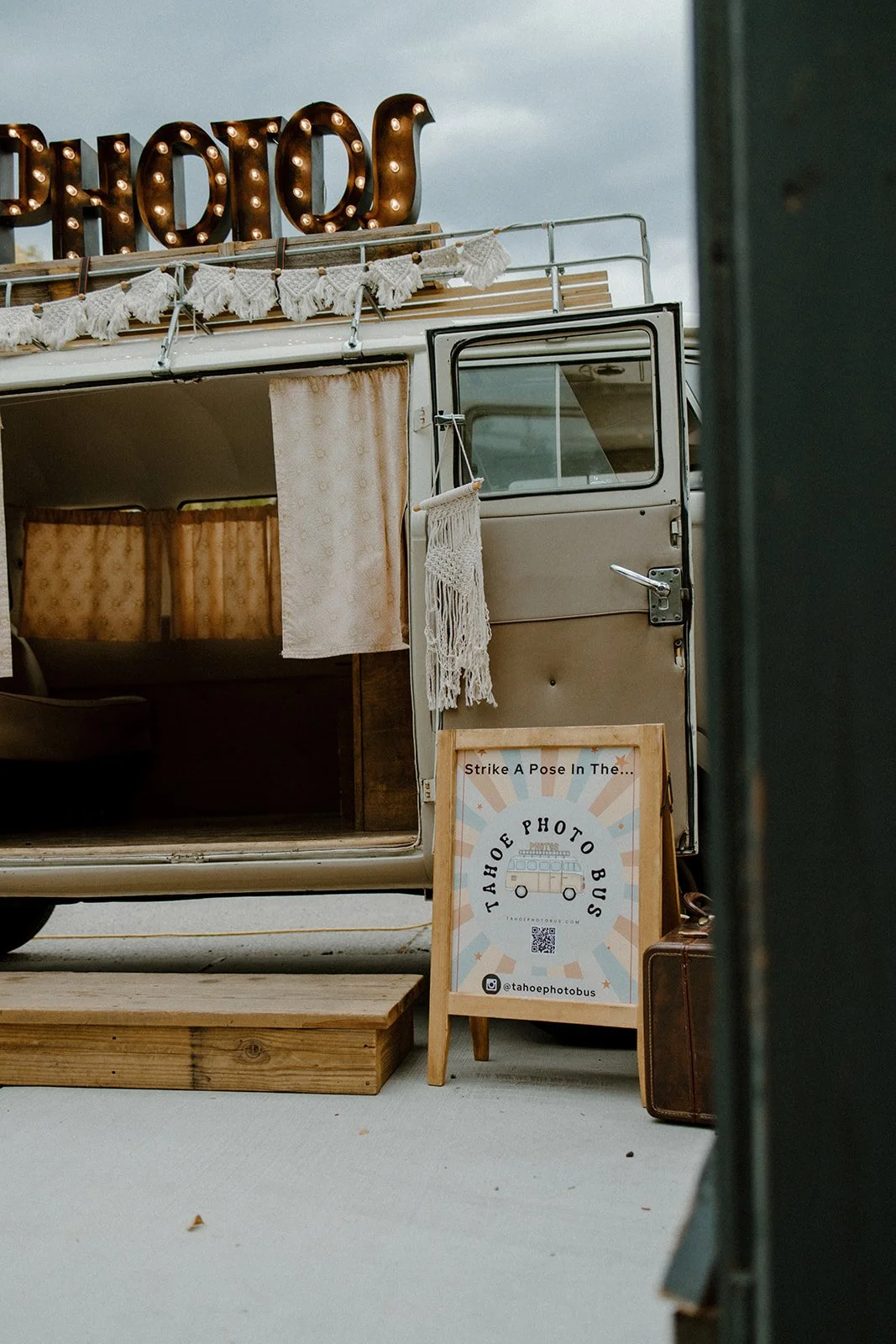 Vintage photo booth in airstream trailer with a sign that reads "Tahoe Photo Bus" and social media handle, decorated with fringe and yarn tassels, outdoors on a cloudy day.