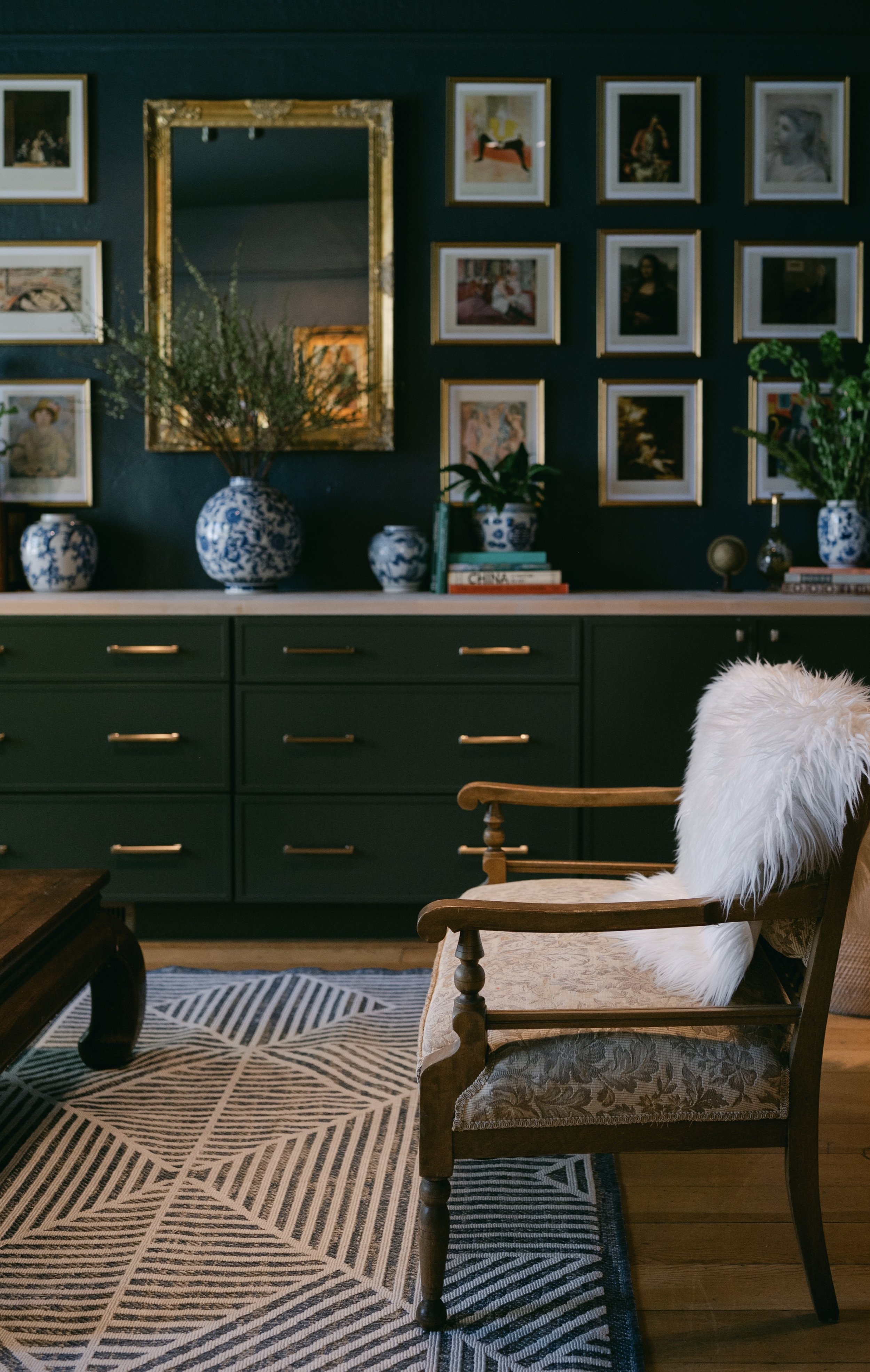 Interior of a room with a dark green sideboard decorated with blue and white vases and a stack of books, a gallery wall with framed artwork, a gold-framed mirror, a wooden armchair with a cushioned seat and feathered white throw, and a patterned blac