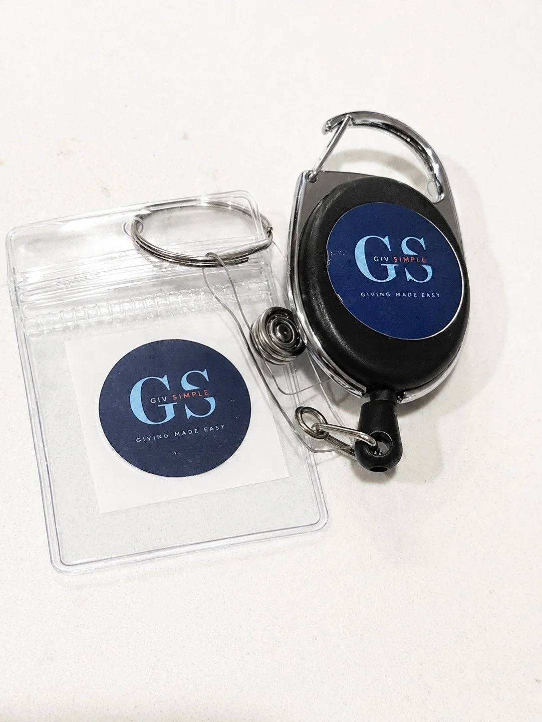 A retractable badge reel with a black casing, metal clip, and blue logo, placed on a white surface.