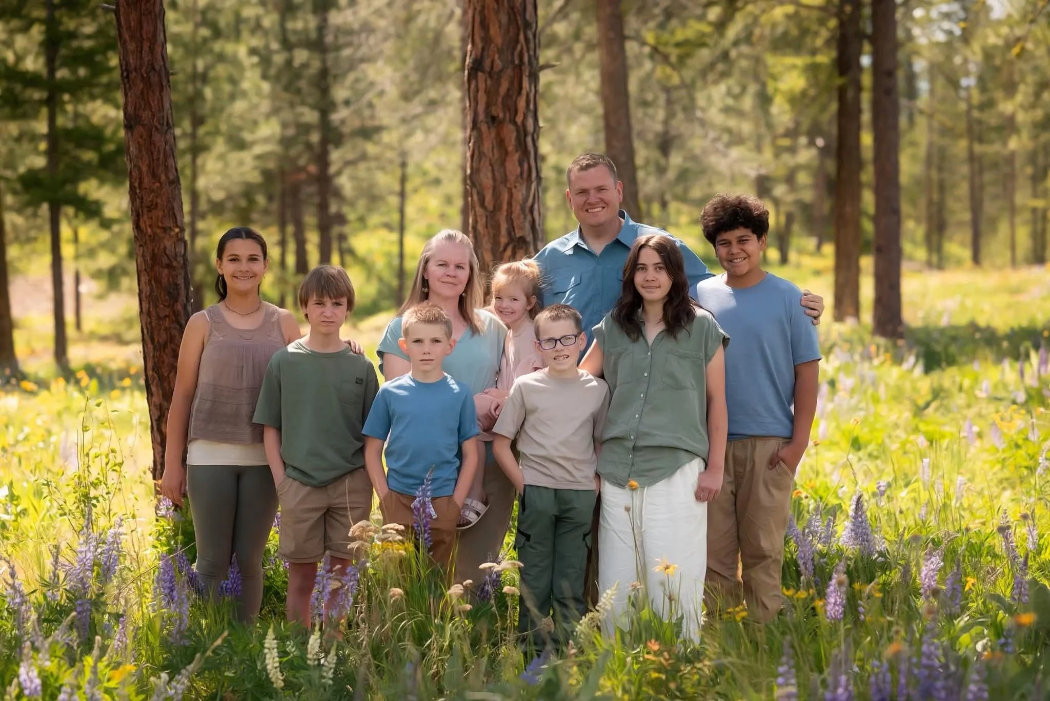 A diverse group of ten people, including children and adults, standing together outdoors in a forested area with trees and wildflowers, smiling at the camera.