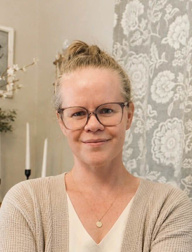 A woman with blonde hair tied in a bun, wearing glasses, a white top, and a beige cardigan, smiling at the camera. She has a gold necklace with a small round pendant. The background features light-colored floral patterned curtains and decor.
