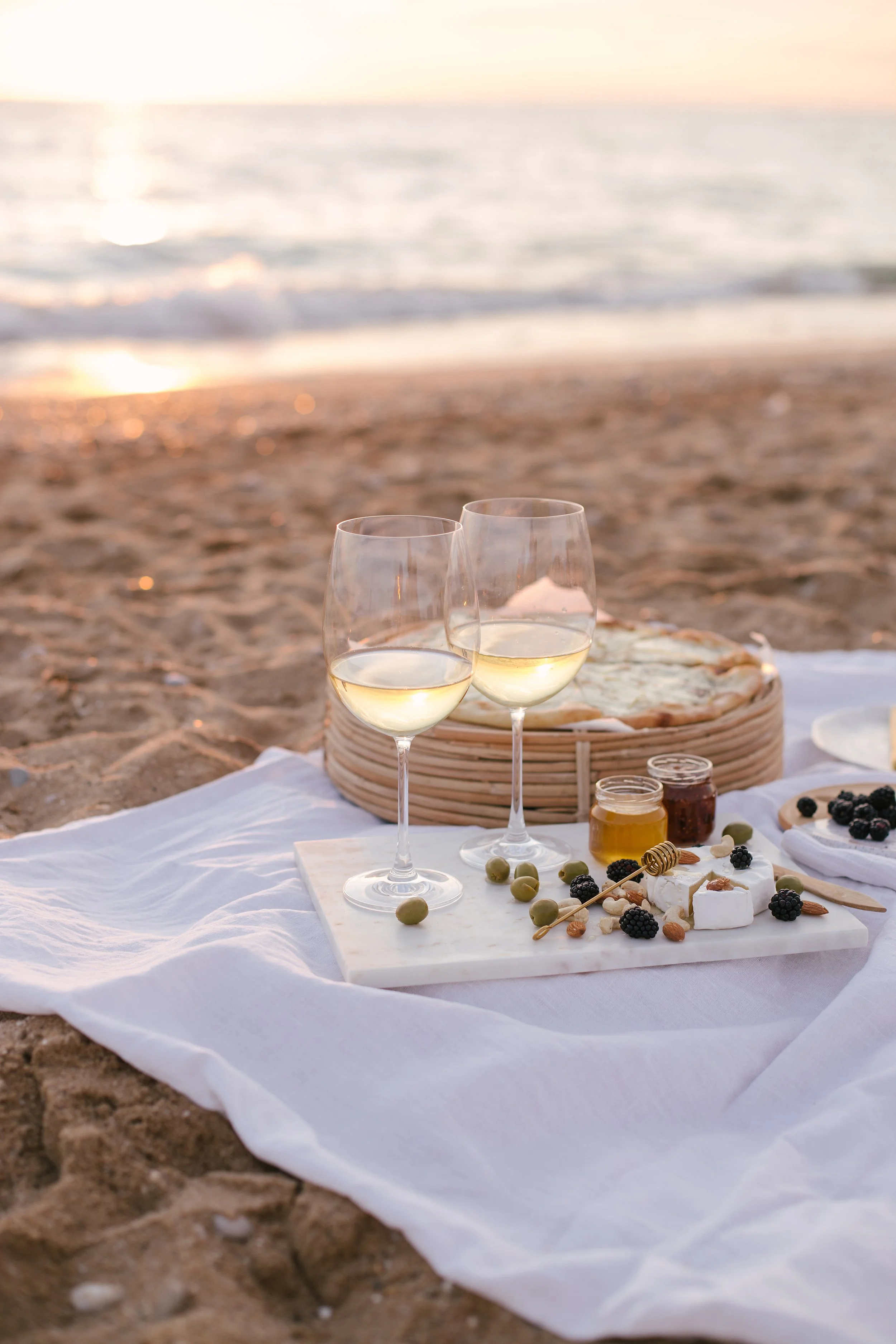 A picnic setup on the beach with two glasses of white wine, a cheese platter with blackberries, green olives, almonds, honey, and a small jar of jam, set on a white cloth with a sunset over the ocean in the background.