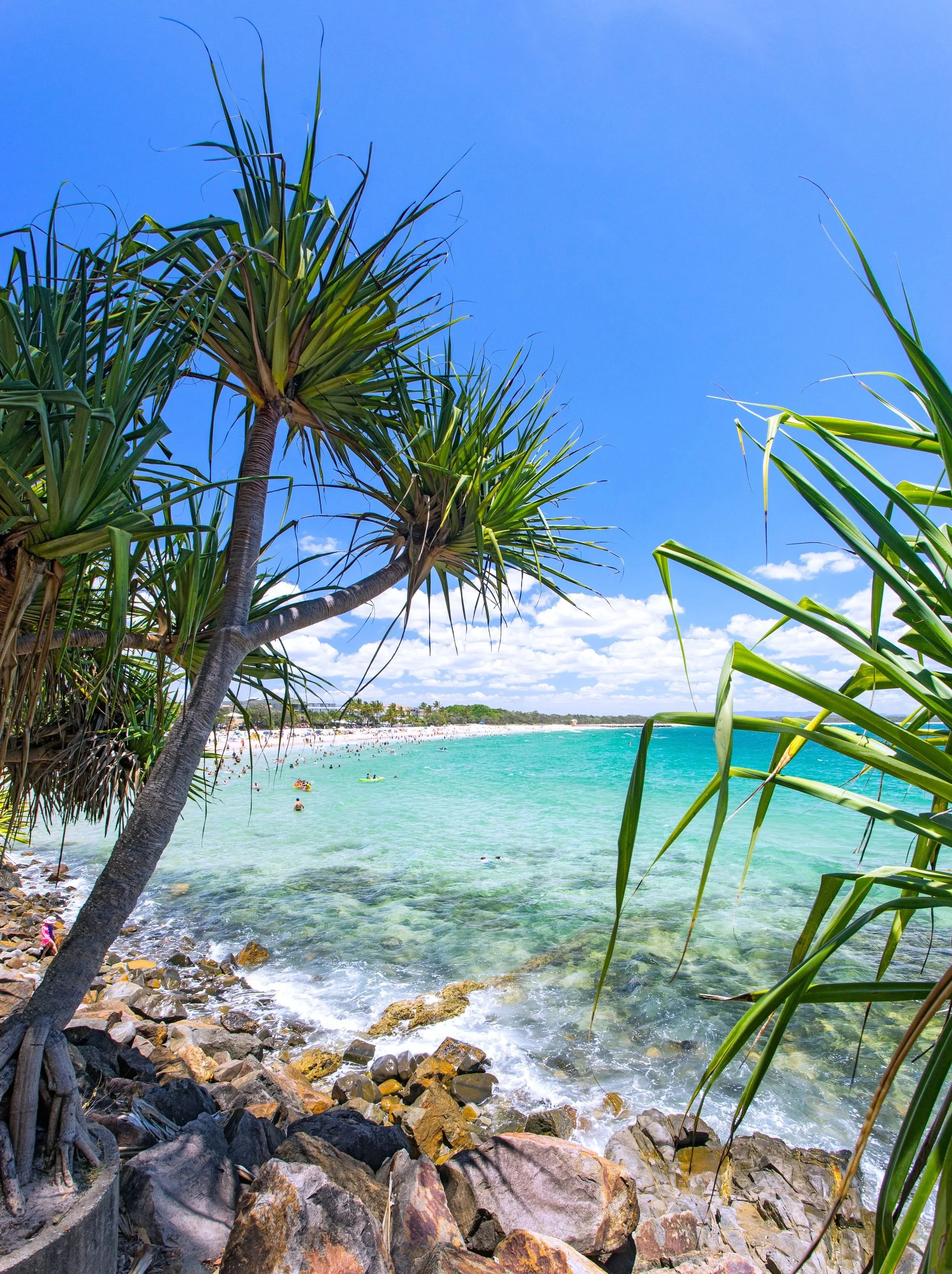 View of a rocky shoreline with clear turquoise water, tropical trees, and a busy beach filled with people, under a bright blue sky with few clouds.