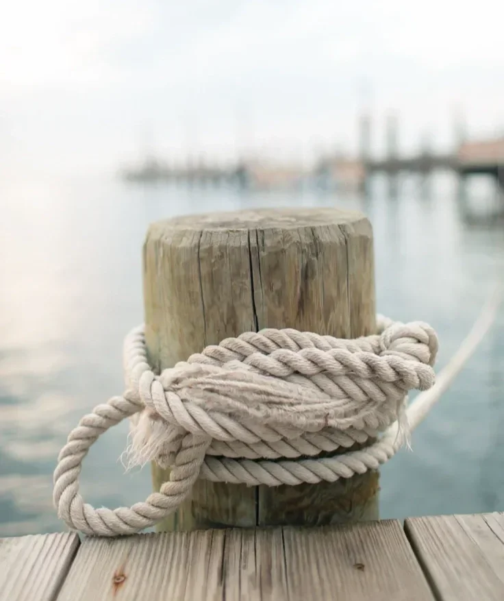 A thick, beige rope tied around a wooden dock post near a body of water, with boats and docks visible in the blurred background.