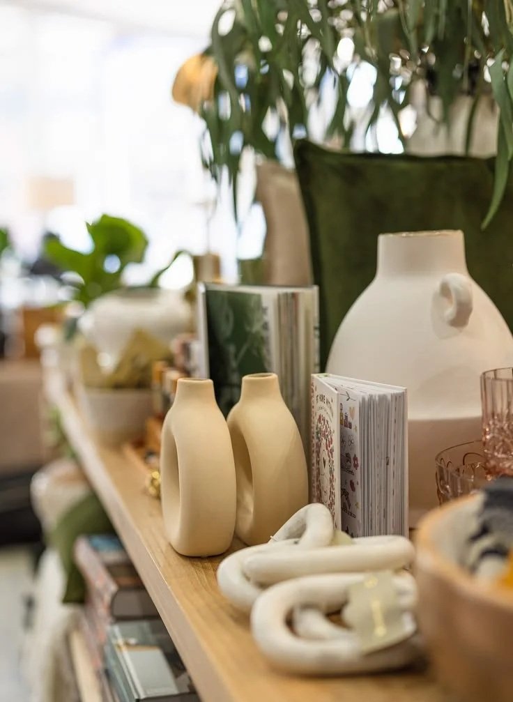 Display of ceramic and glass vases, books, and decorative objects on a wooden shelf with plants in the background.