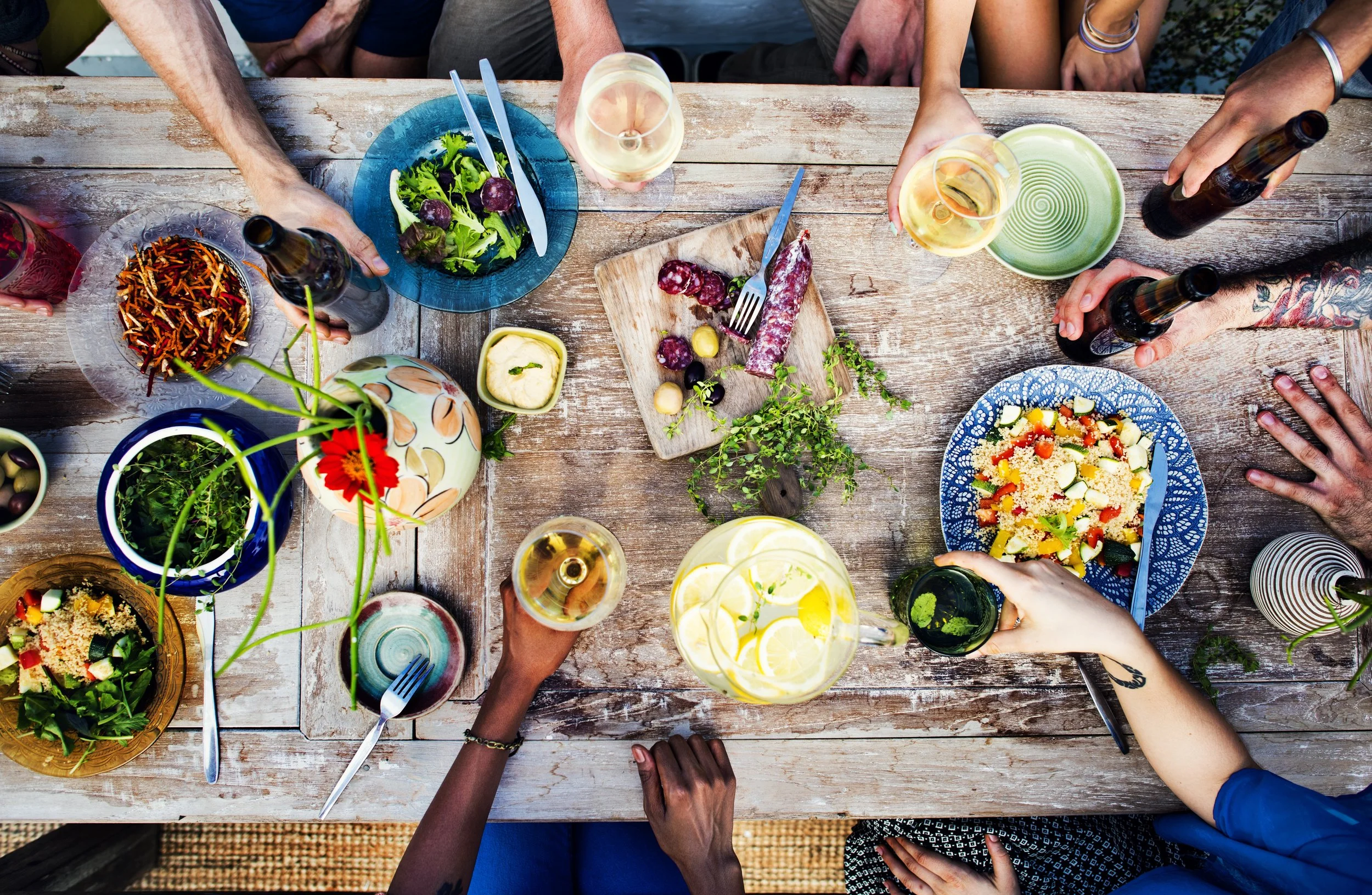 People sitting around a rustic wooden table enjoying a meal with various salads, drinks, and a wooden cutting board with sausages and grapes.