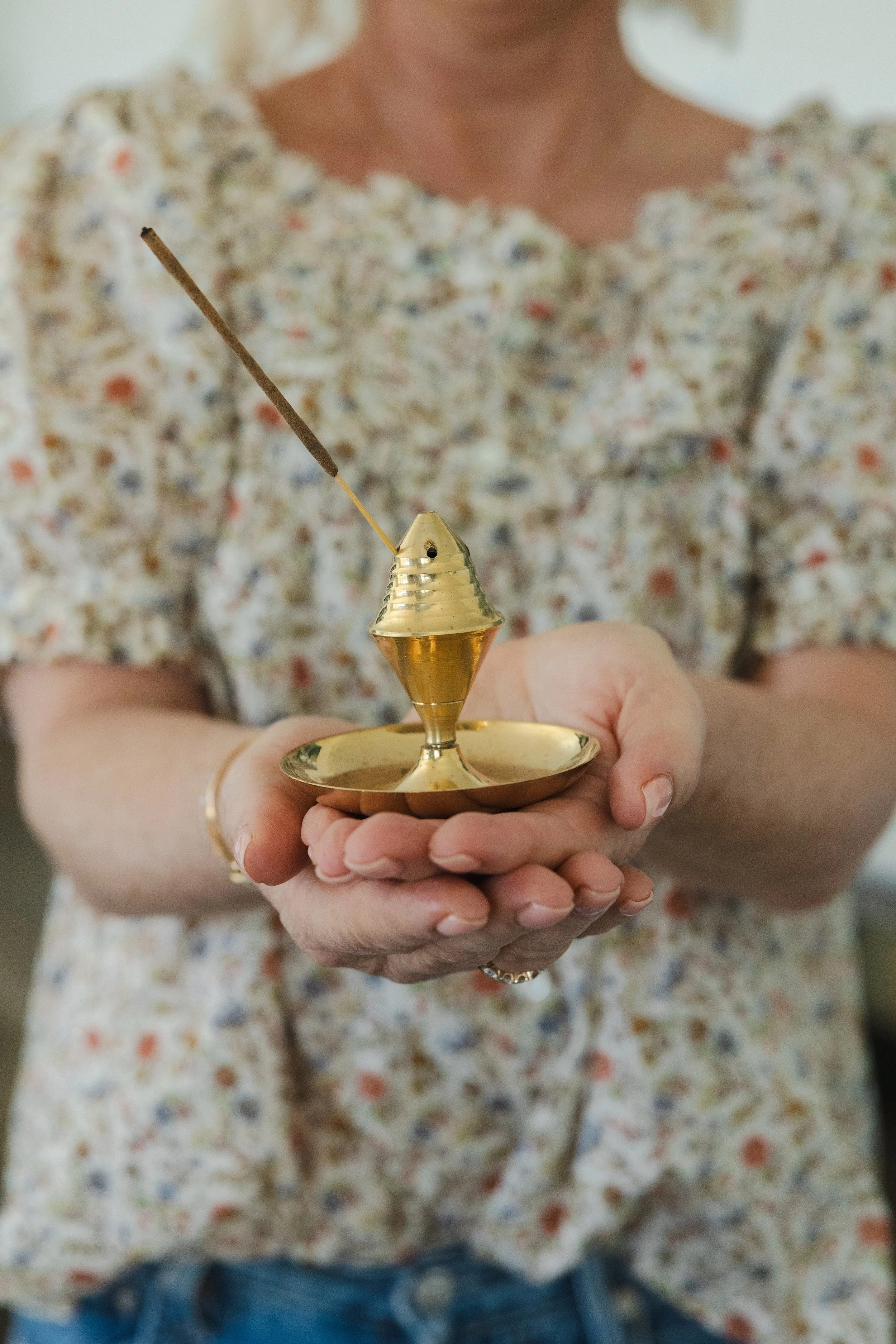 A person holding a small, golden incense holder with a stick of incense burning inside it.