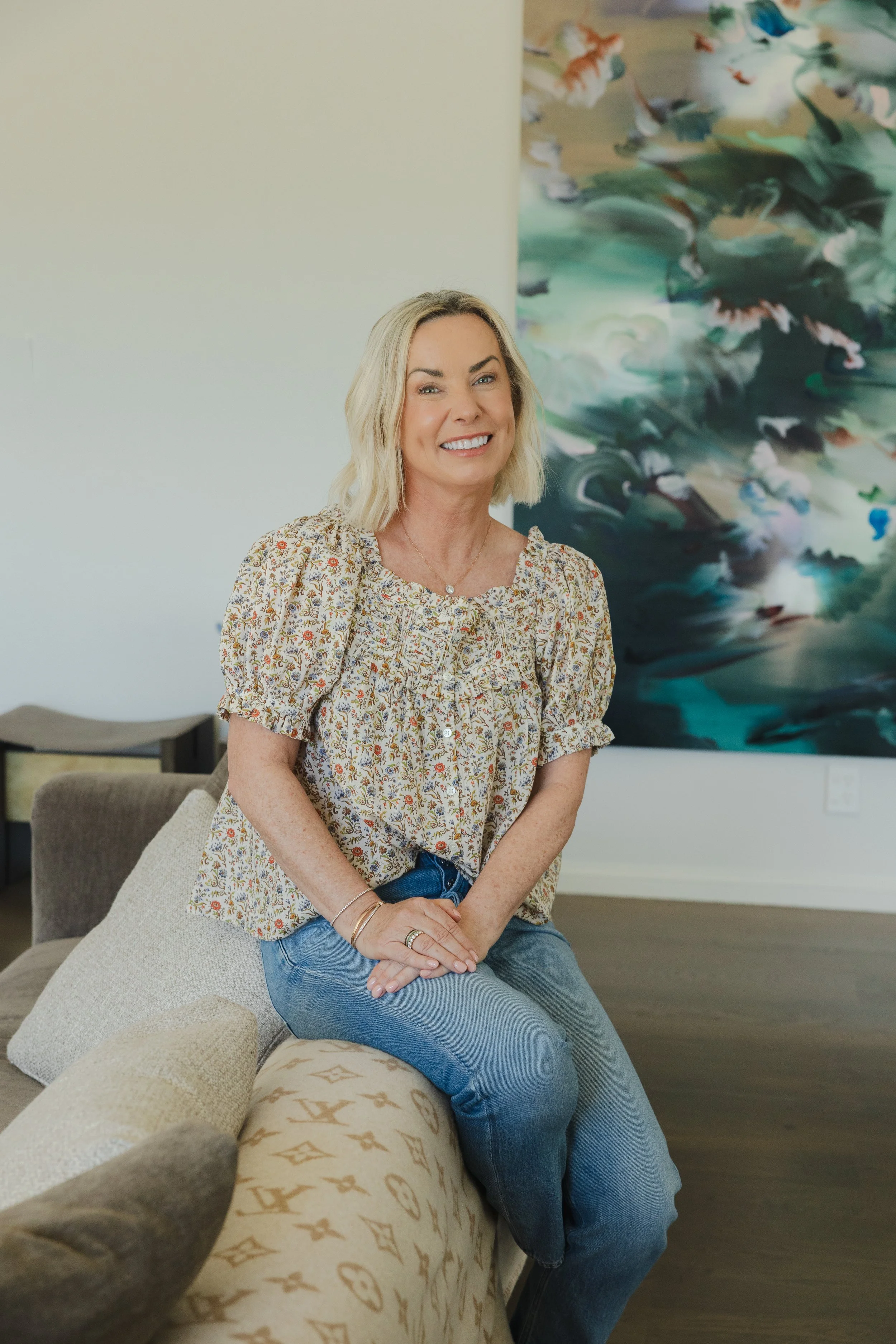 A woman with blonde hair, wearing a floral blouse and jeans, sitting on a beige patterned sofa in a modern room with abstract artwork on the wall behind her, smiling at the camera.