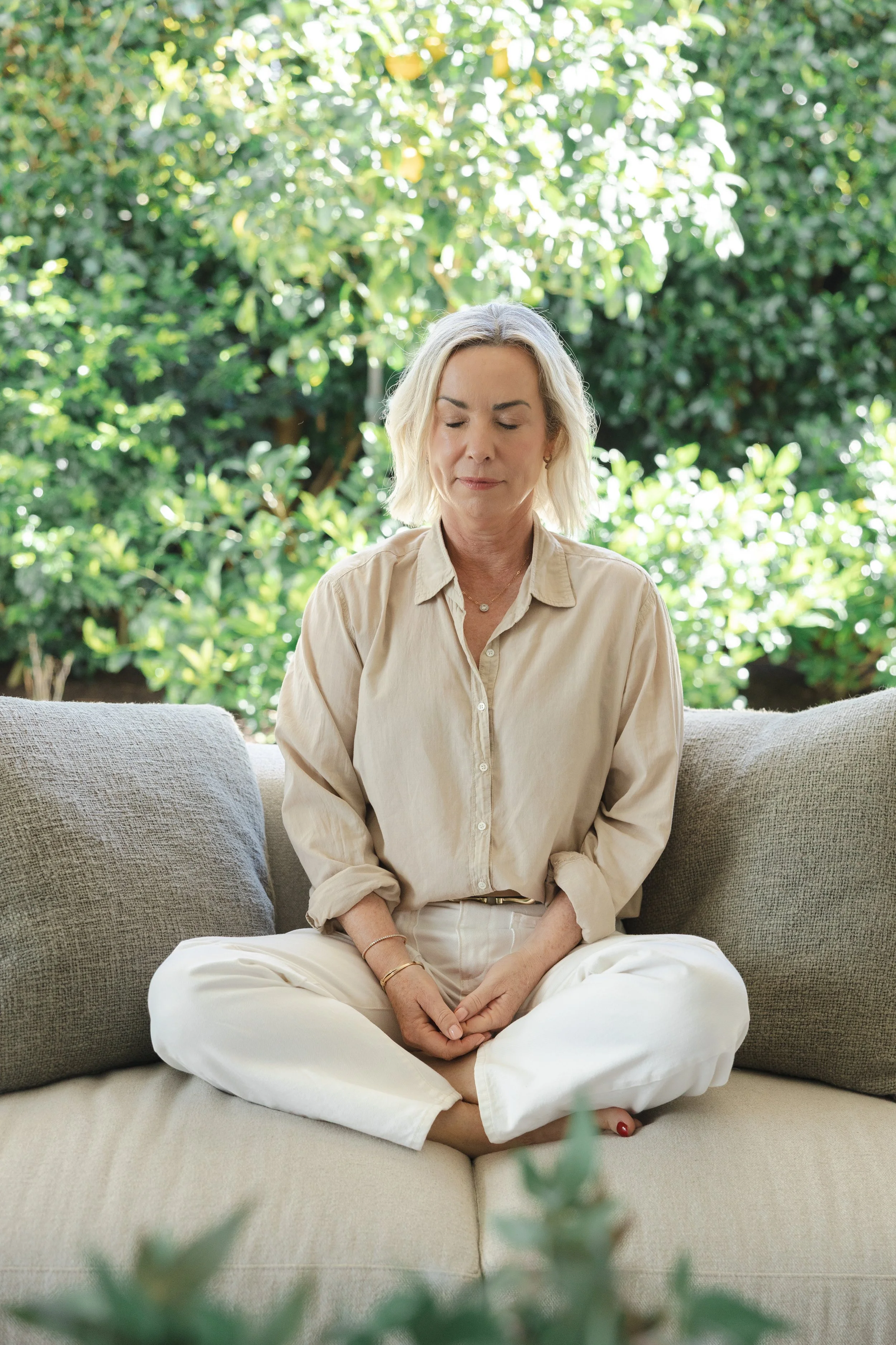 A woman with blonde hair sitting cross-legged on a beige sofa with her eyes closed, surrounded by green foliage outside in natural light.
