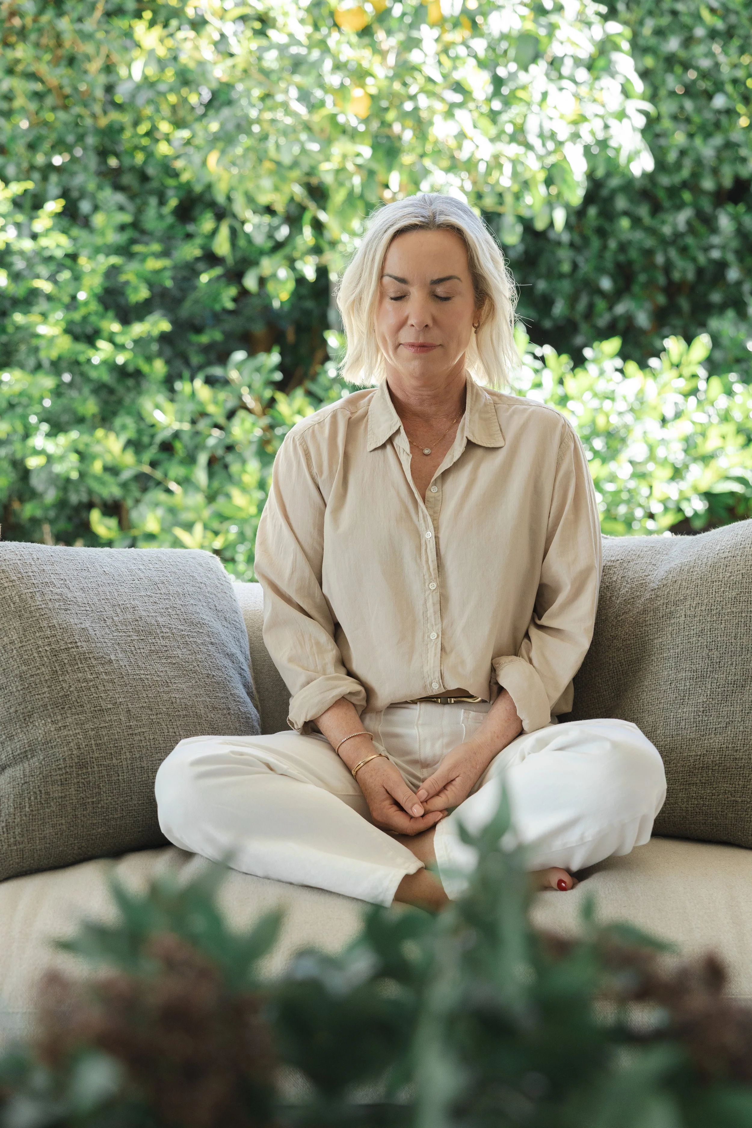 A woman with blonde hair sitting cross-legged on a sofa with closed eyes, meditating or relaxing in a bright outdoor setting with trees in the background.