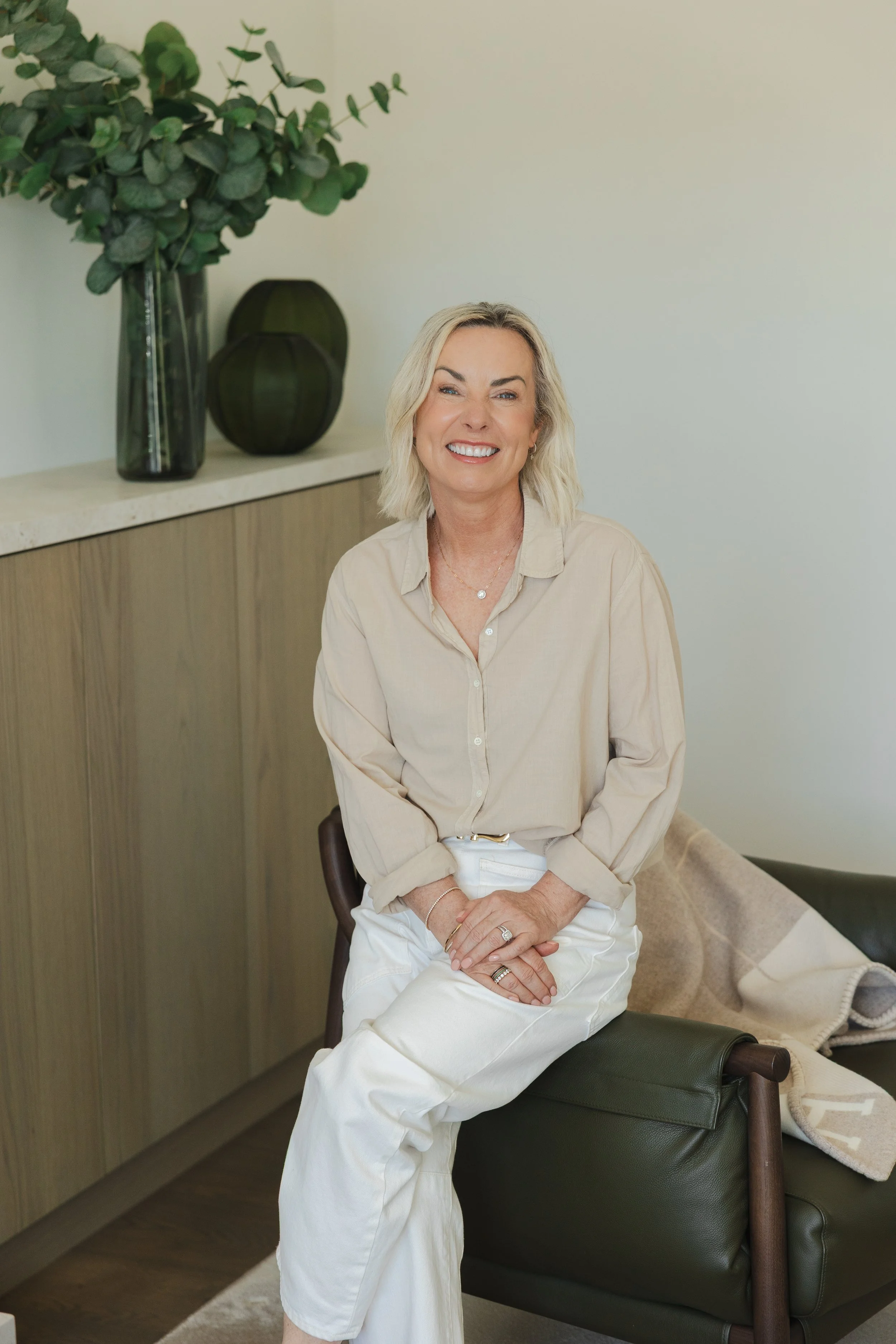 A smiling middle-aged woman with blonde hair, wearing a beige button-up shirt and white pants, sitting on a bench in a modern, minimalist room with green and black decorative vases and a large leafy plant in the background.