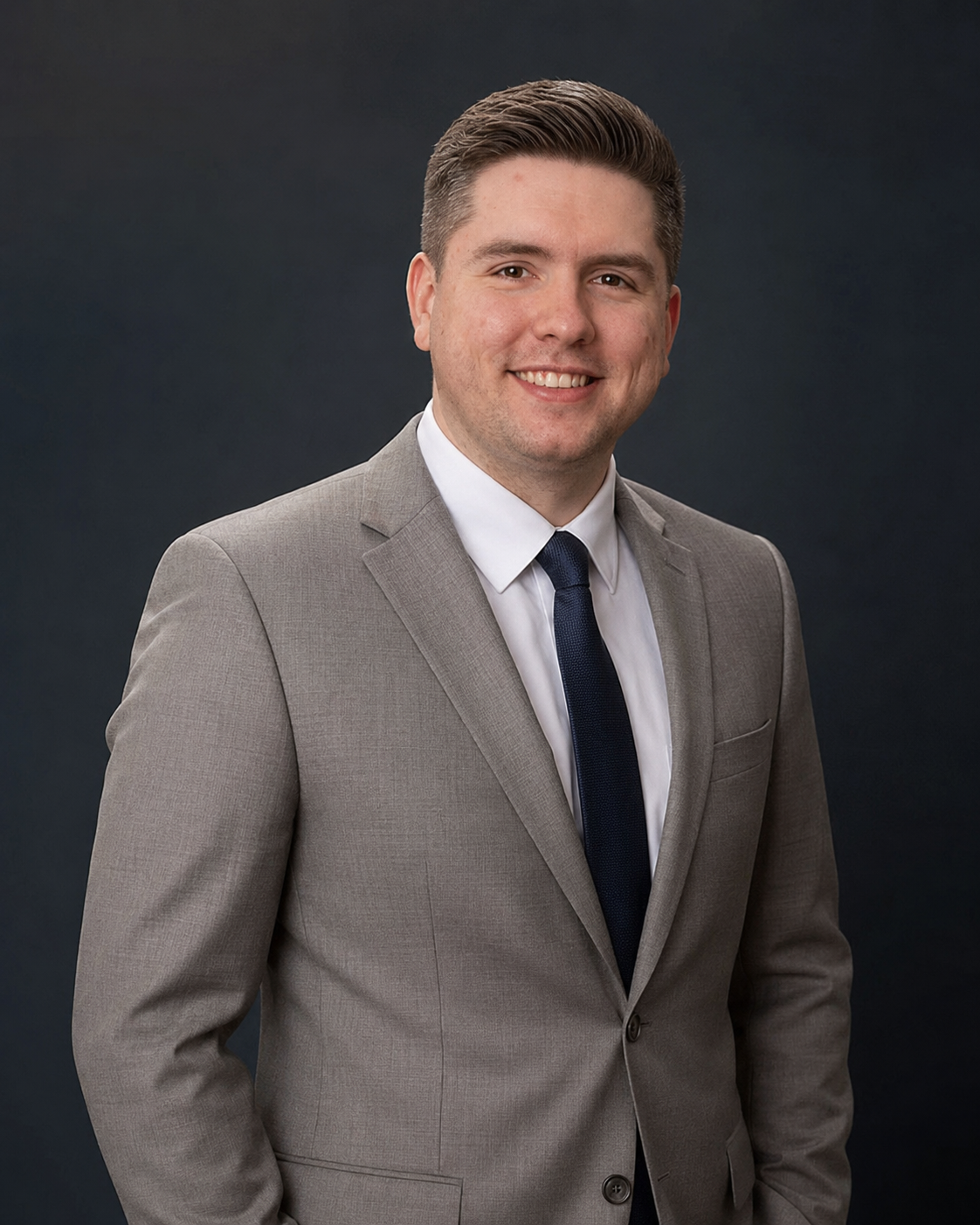 Professional young man in gray suit, white shirt, navy tie, smiling against dark background.