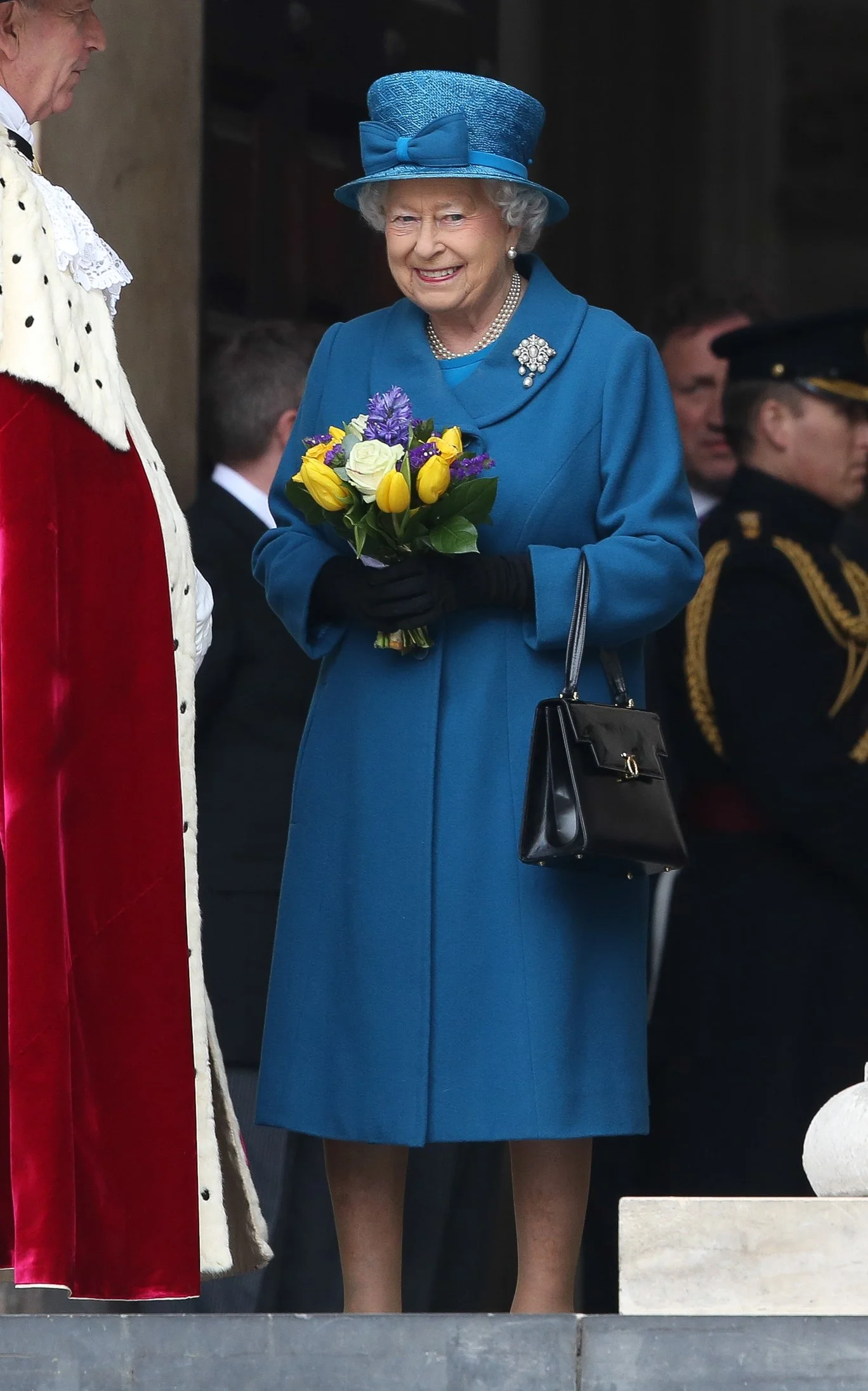 Queen Elizabeth II wearing a blue coat, matching hat, pearl necklace, and brooch, holding a bouquet of flowers.