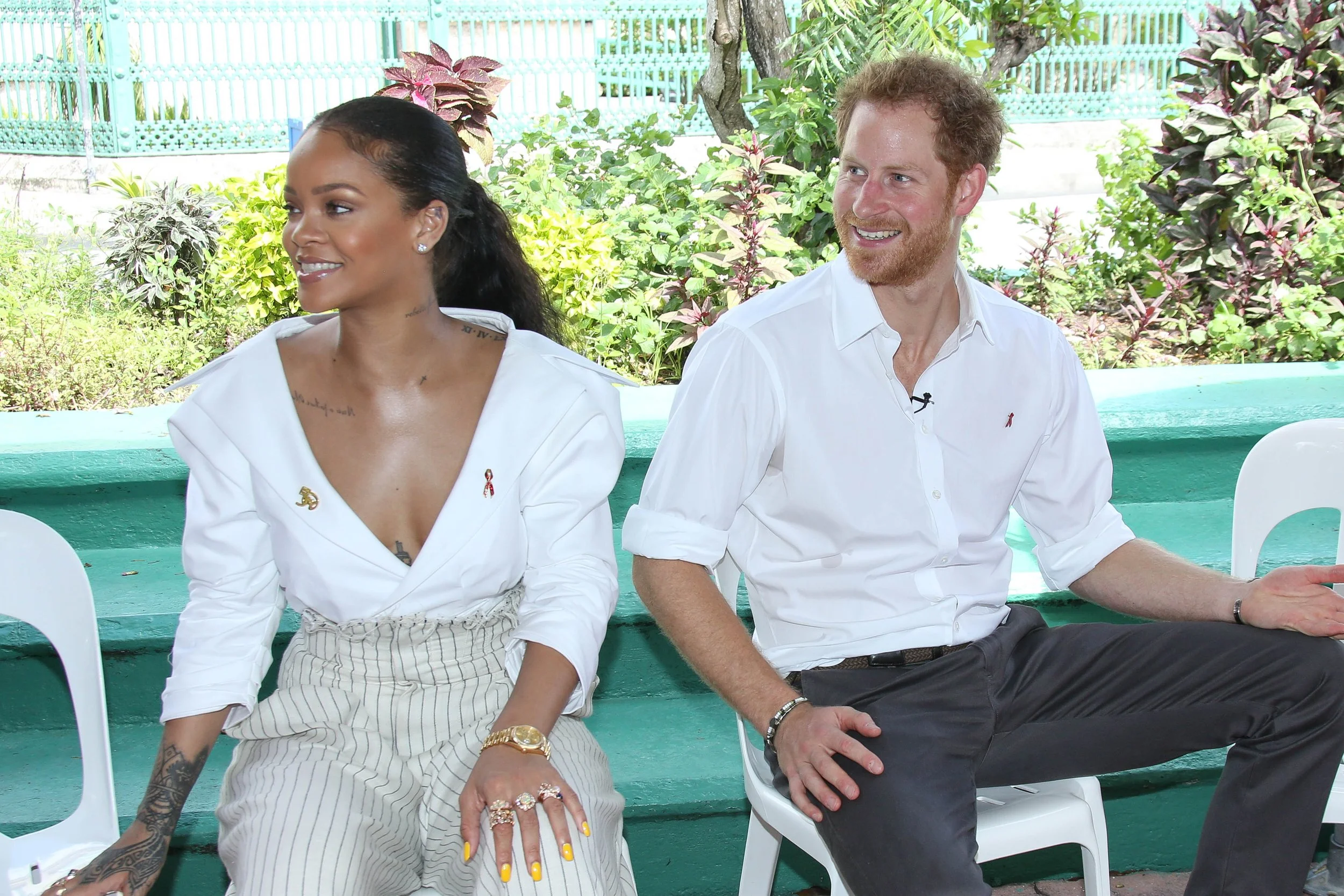 A woman with dark hair in a ponytail and tattoos on her arm, wearing a white shirt, seated next to a man with curly red hair and beard, wearing a white shirt, both smiling and sitting outdoors with greenery in the background.