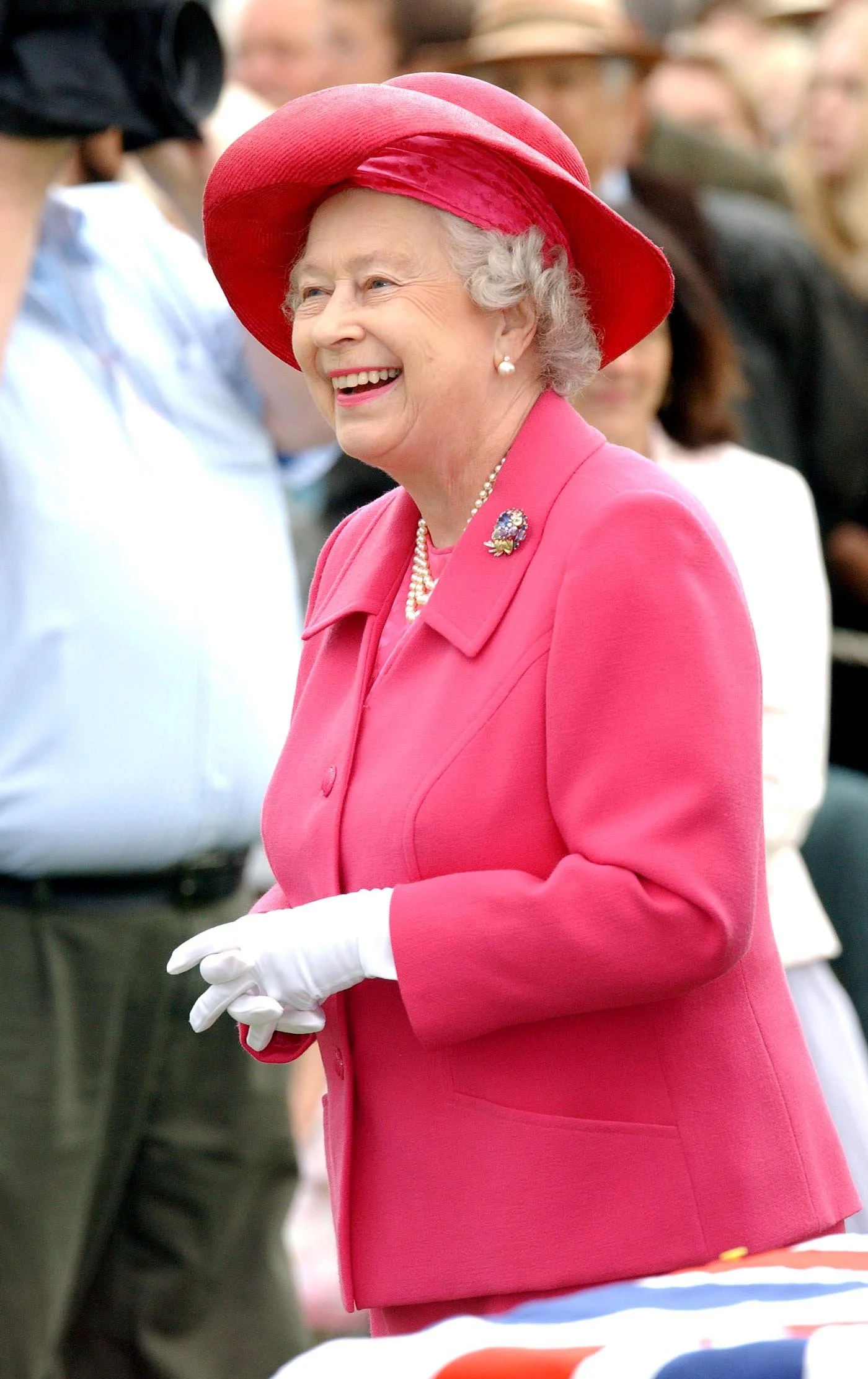 Queen Elizabeth II smiling, wearing a pink coat, a red hat, white gloves, pearl earrings, and a brooch. Guards Polo Club Berkshire