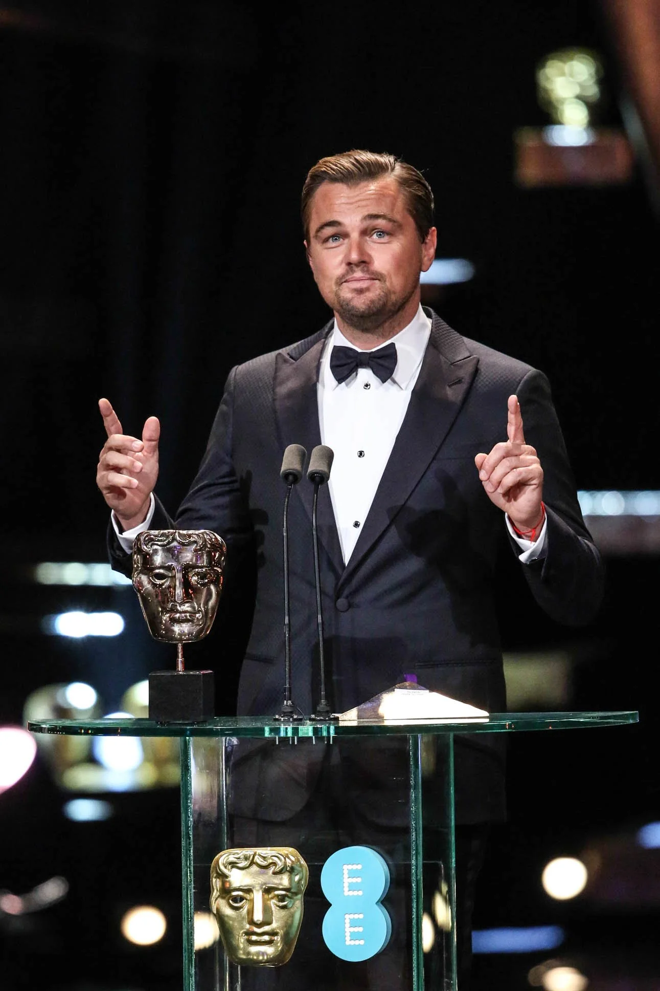 Actor Leonardo DiCaprio in a tuxedo standing behind an awards podium, giving a speech at an awards ceremony, with BAFTA trophies and a glass table in front of him.