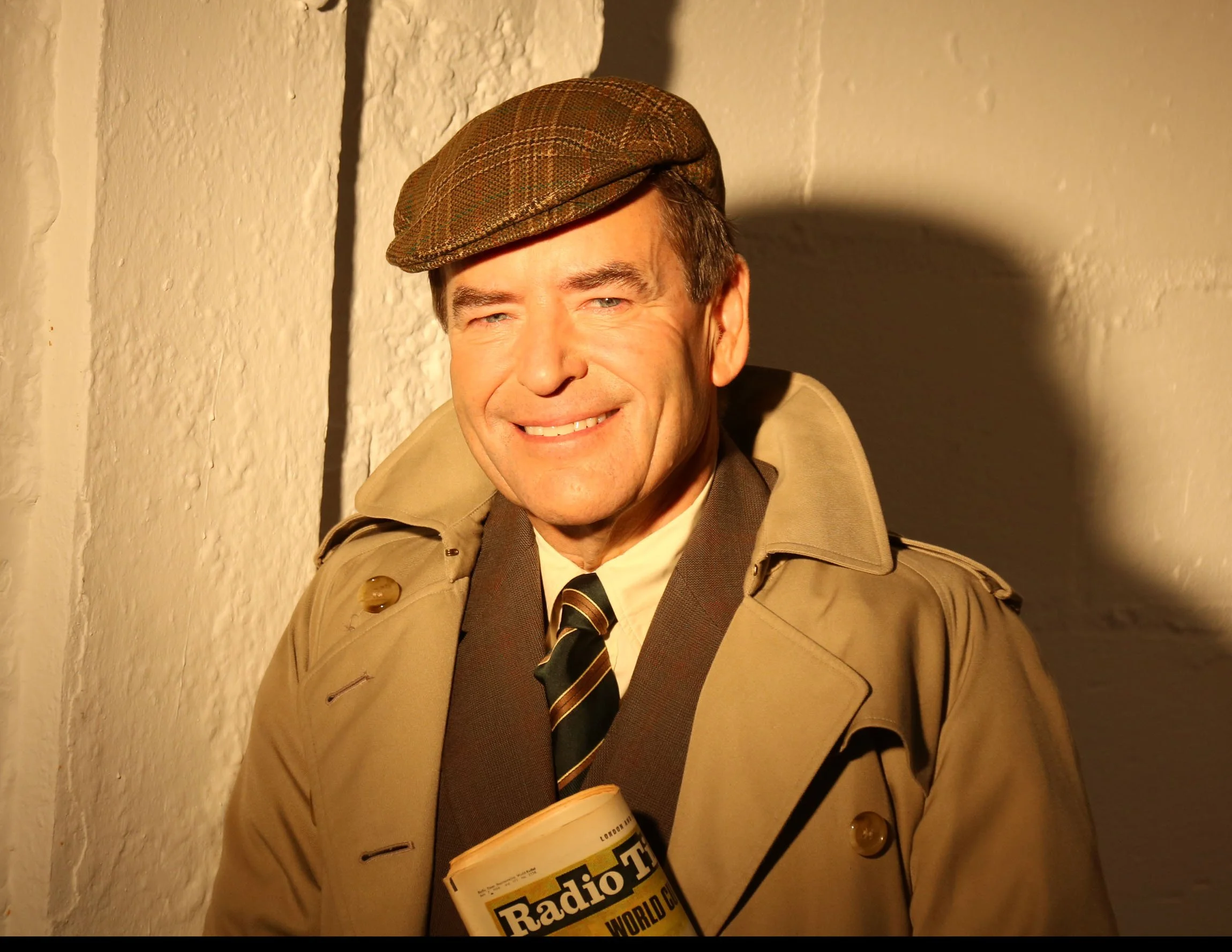 A man wearing a brown plaid flat cap, beige trench coat, and black and yellow striped tie, smiling while holding a cup and a newspaper titled "Radio" against a textured beige wall.