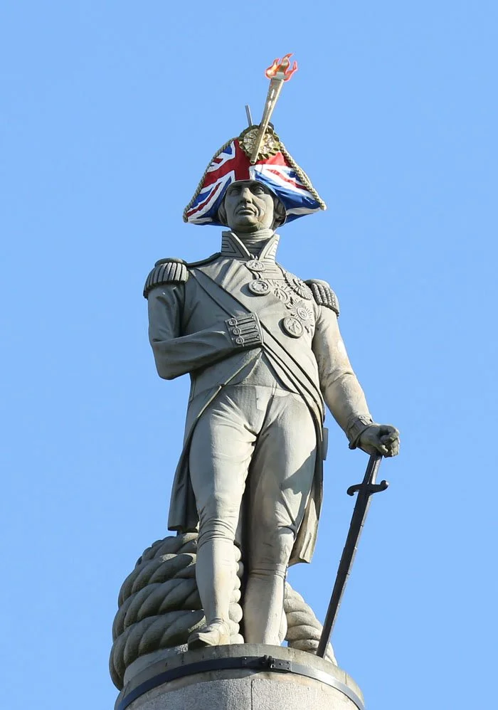 Statue of a historical military figure wearing a tri-corner hat decorated with a British flag and a torch with flames, holding a sword, against a clear blue sky.