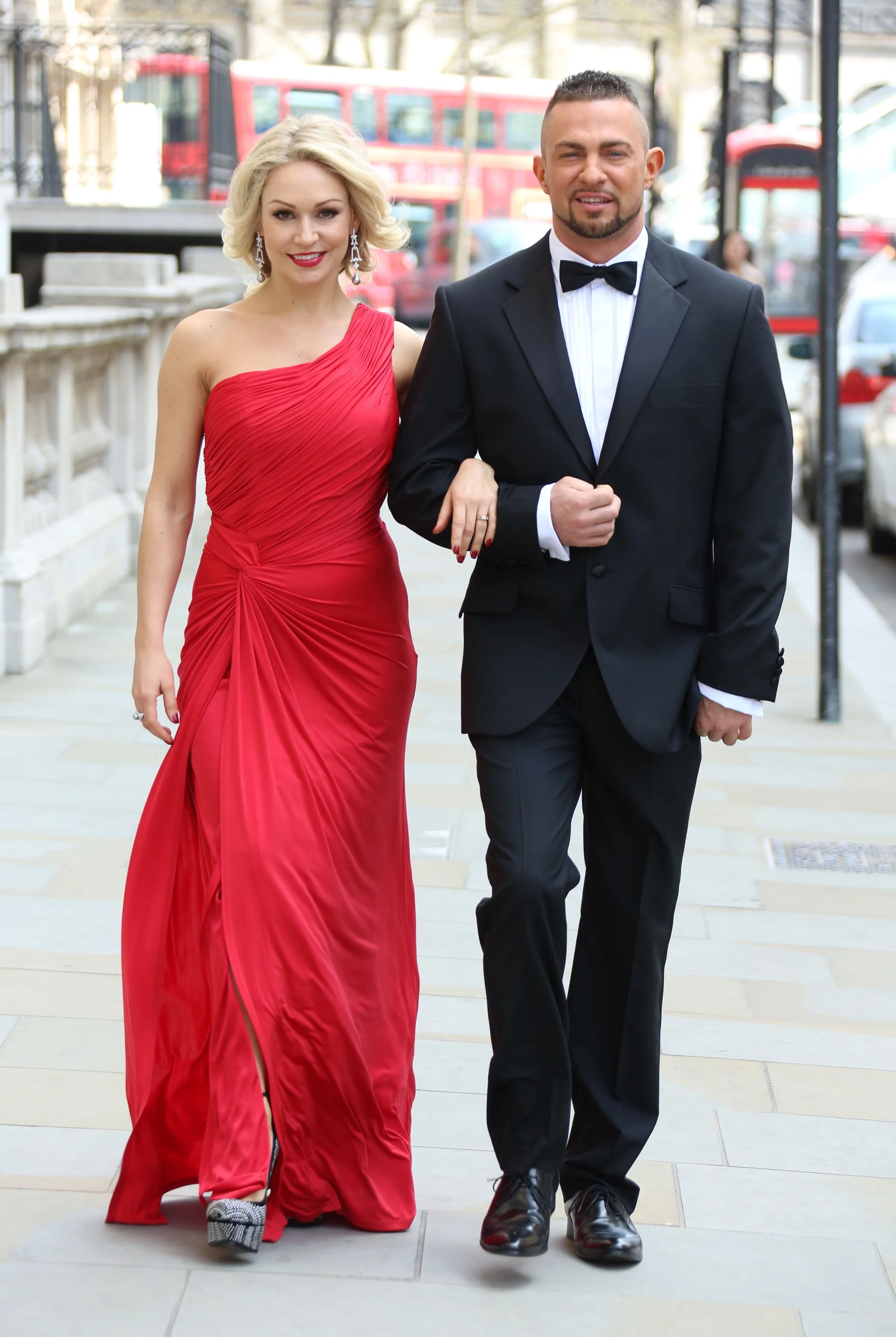 A couple walking on a city sidewalk, the woman in a red gown and the man in a black tuxedo, holding each other arm in arm.