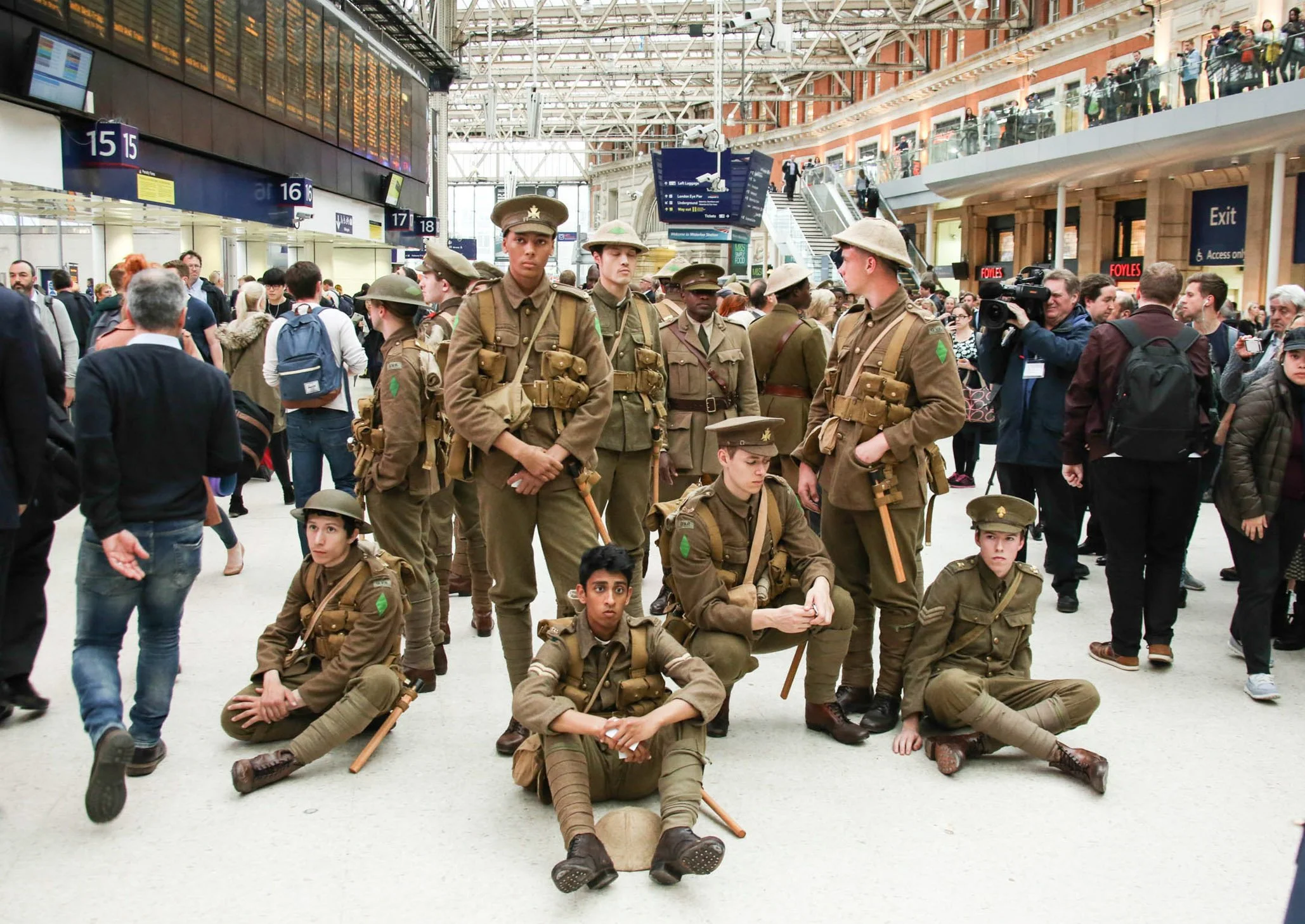 A group of people dressed in military uniforms posing at a busy train station, with other travelers and large digital departure boards in the background.