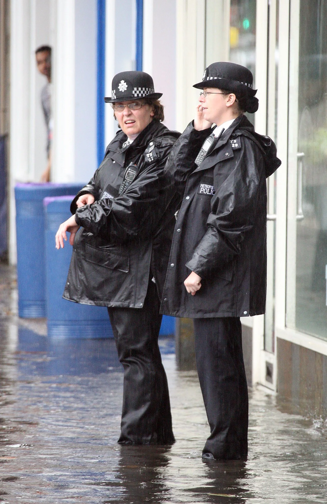 Two police officers standing in a flooded area outside a building, with one talking on a cellphone, wearing black police uniforms and hats.