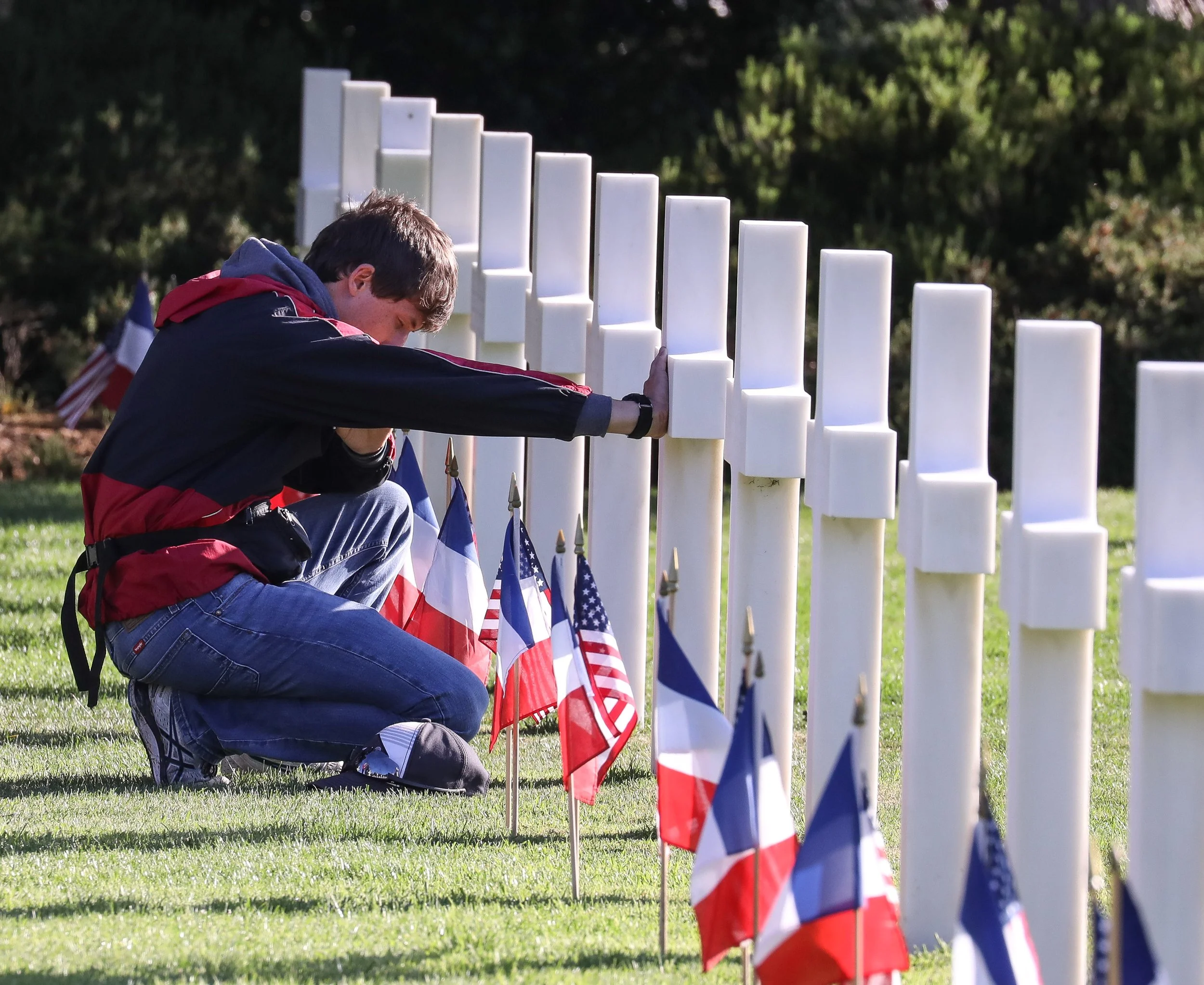 A man kneeling and placing flags near memorial white crosses outdoors during daylight, with dark green trees in the background.