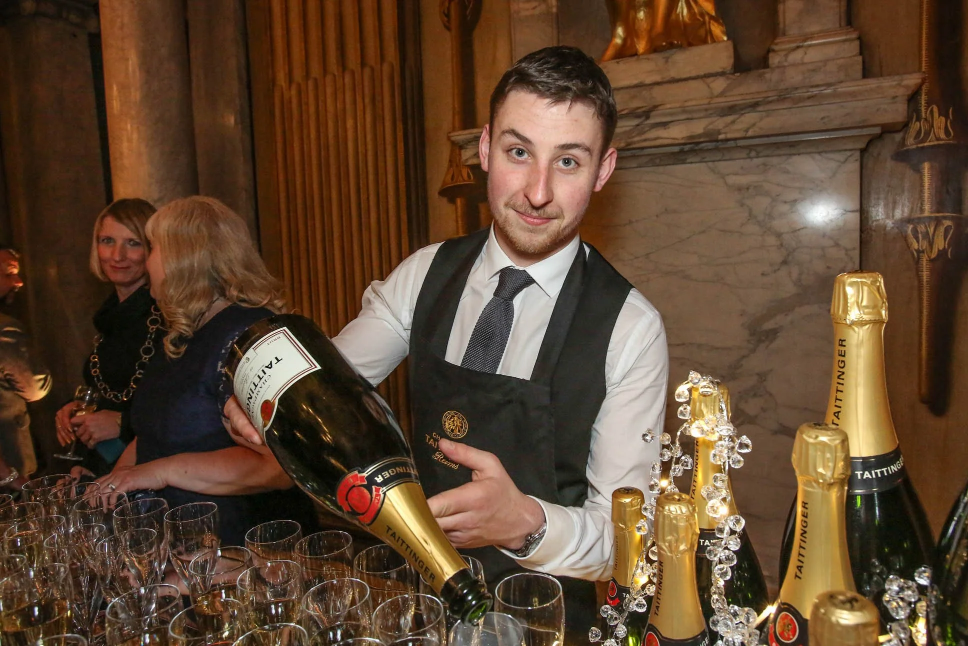 A young man in a white shirt, black tie, and black apron pours champagne from a large bottle into glasses at a celebration event. Behind him, a woman with blonde hair and a dark outfit smiles, with a marble wall and golden decorations in the backgrou