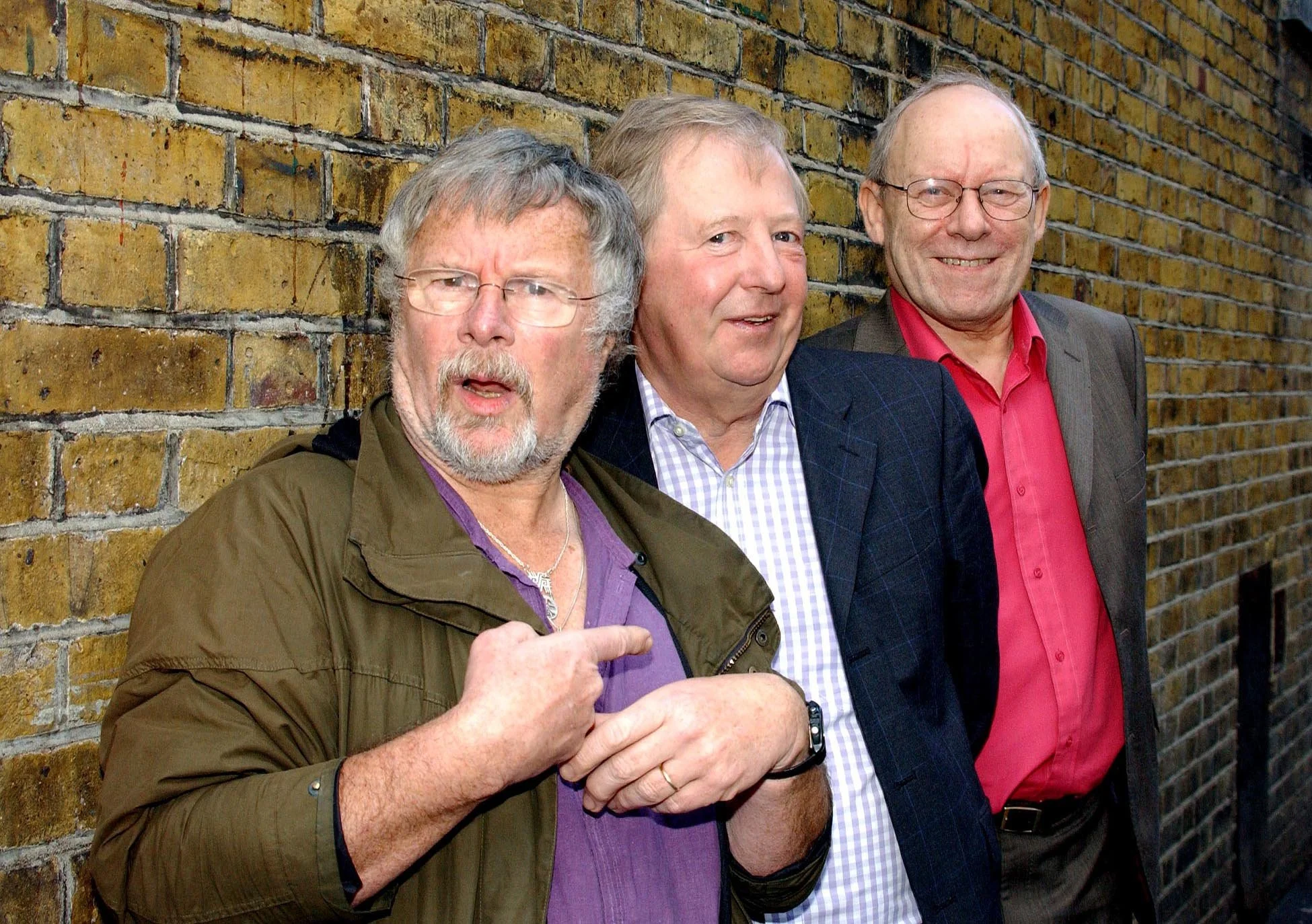 Three middle-aged men standing against a brick wall, smiling, with one man in front gesturing with his hand.