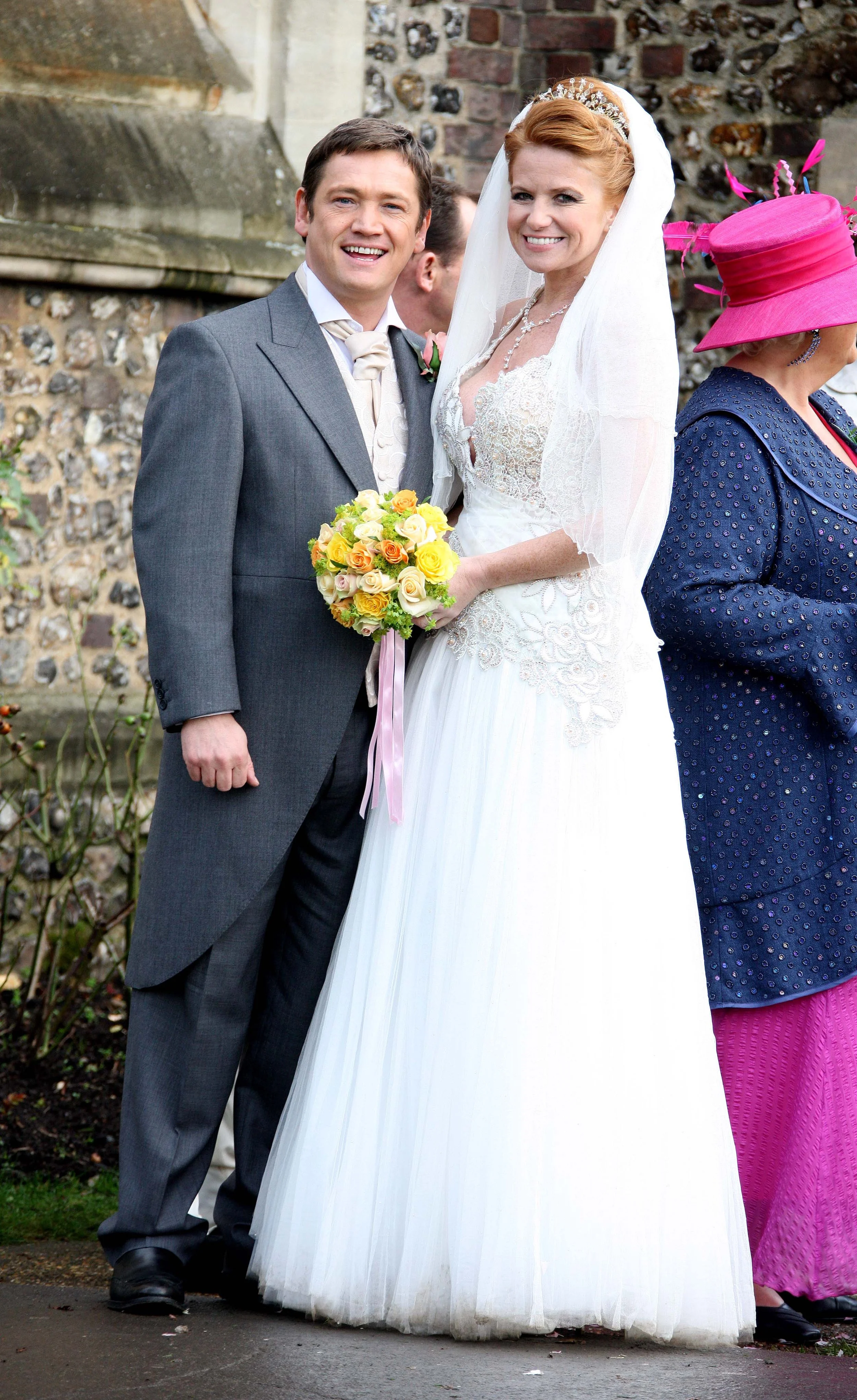 A newlywed couple smiling for a photo outside a church, with a woman in a pink hat and blue jacket visible on the right.