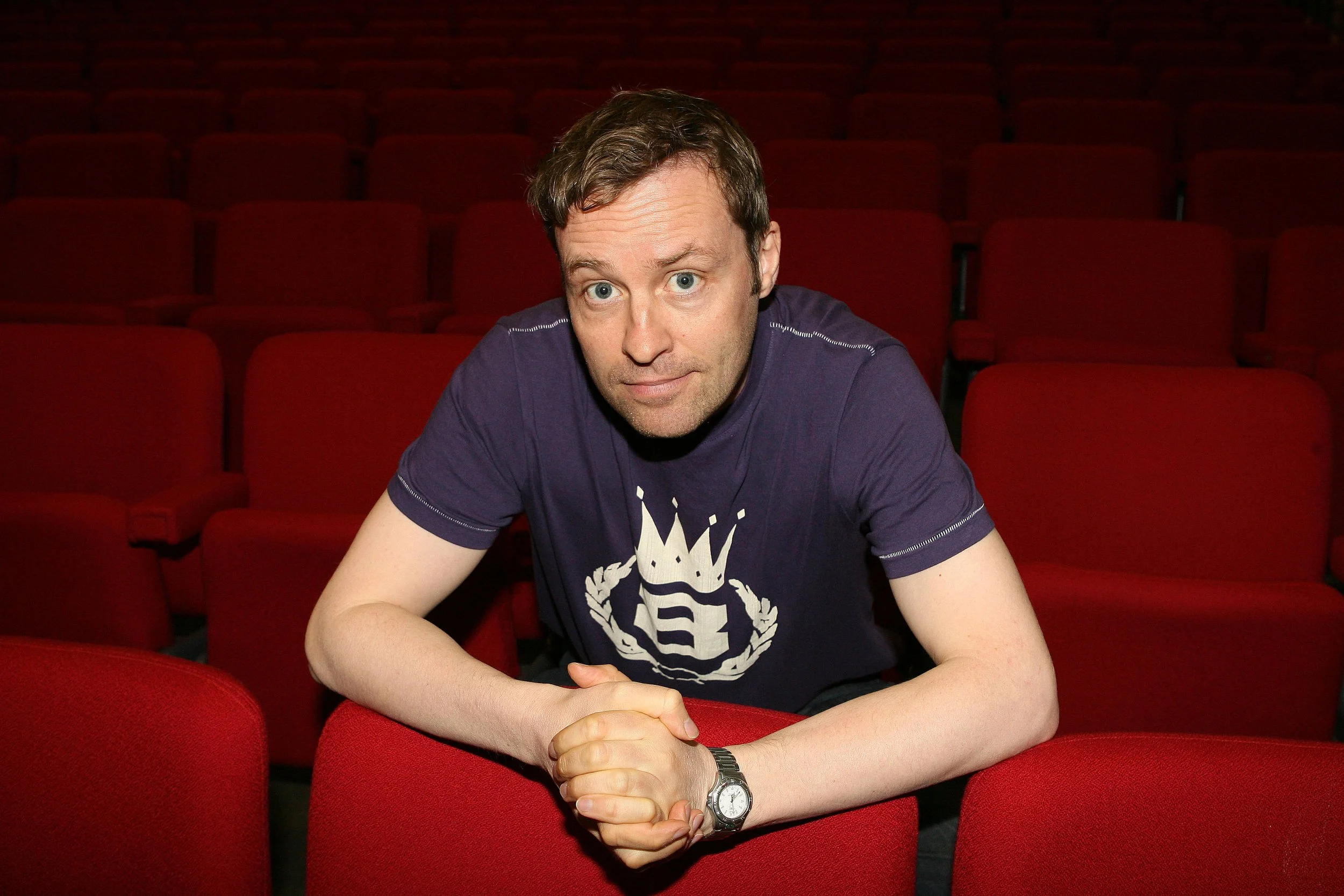 A man with short brown hair and blue eyes sitting in a red theater with empty red seats, wearing a navy blue t-shirt with white graphics, a silver wristwatch, looking at the camera.