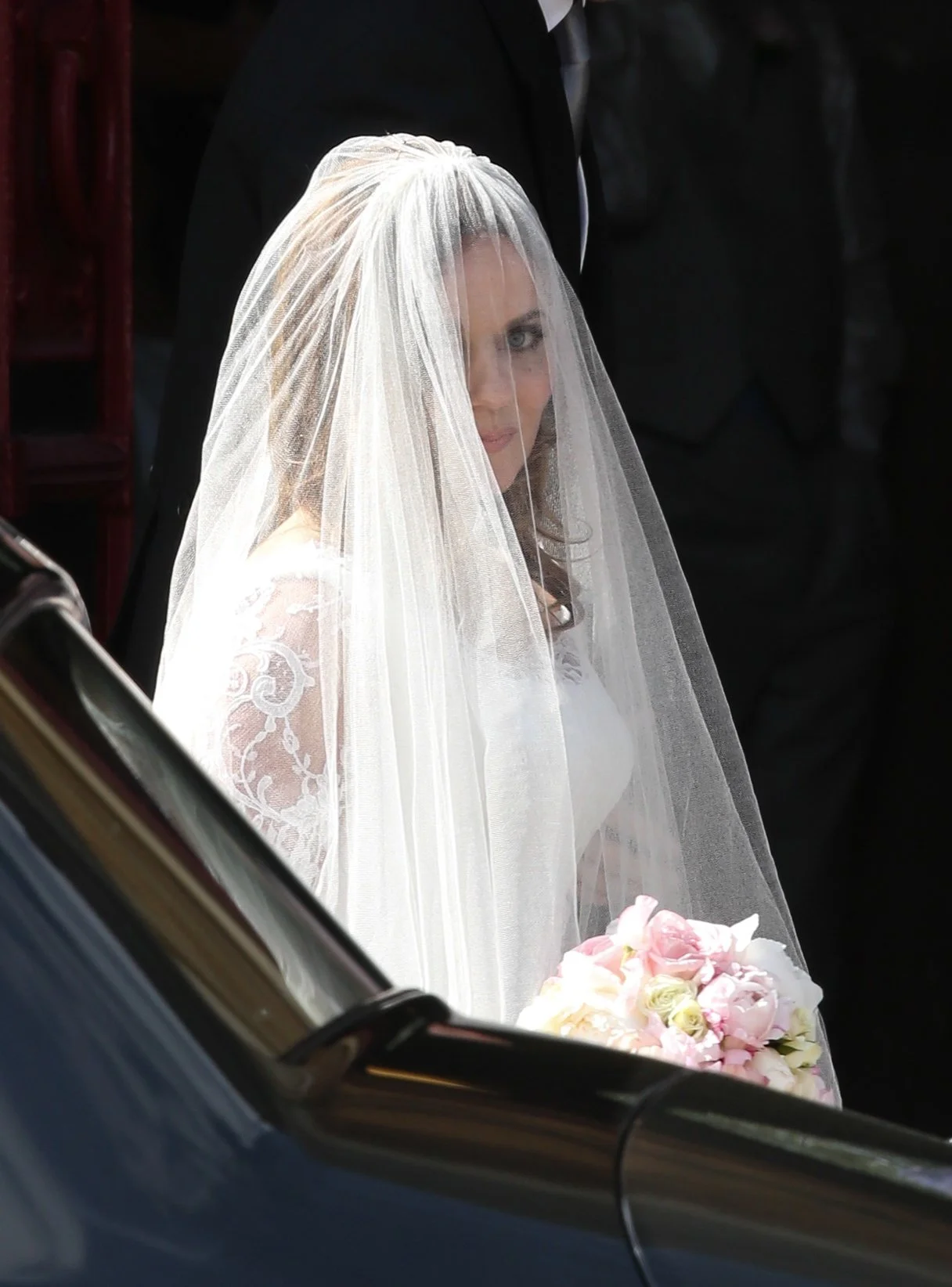 Bride with a veil and white wedding dress holding a bouquet of pink and white flowers, sitting in a car before her wedding ceremony.
