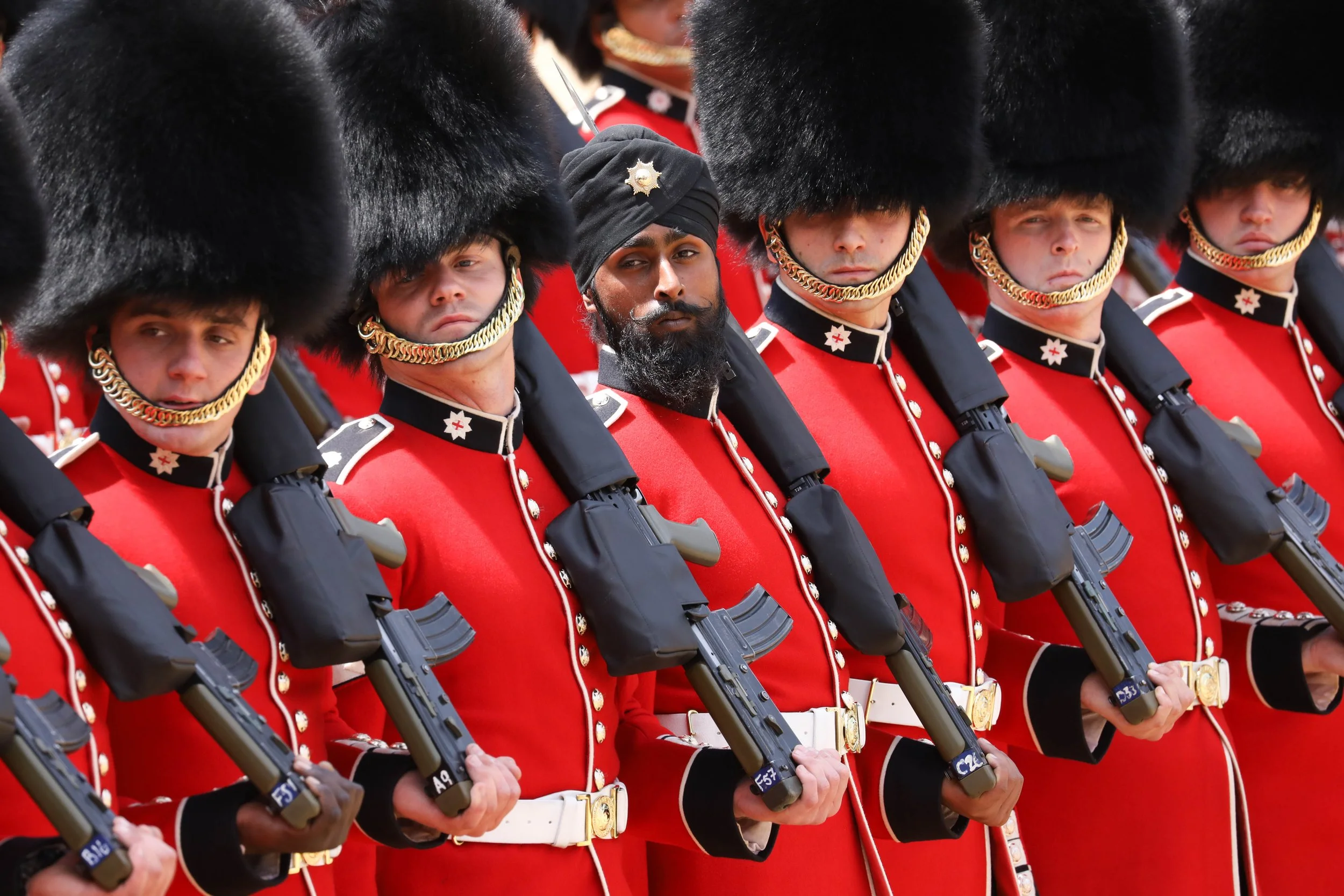 A group of soldiers dressed in red uniforms with black tall fur hats, gold chains, and holding rifles, standing in a line during a ceremonial event.