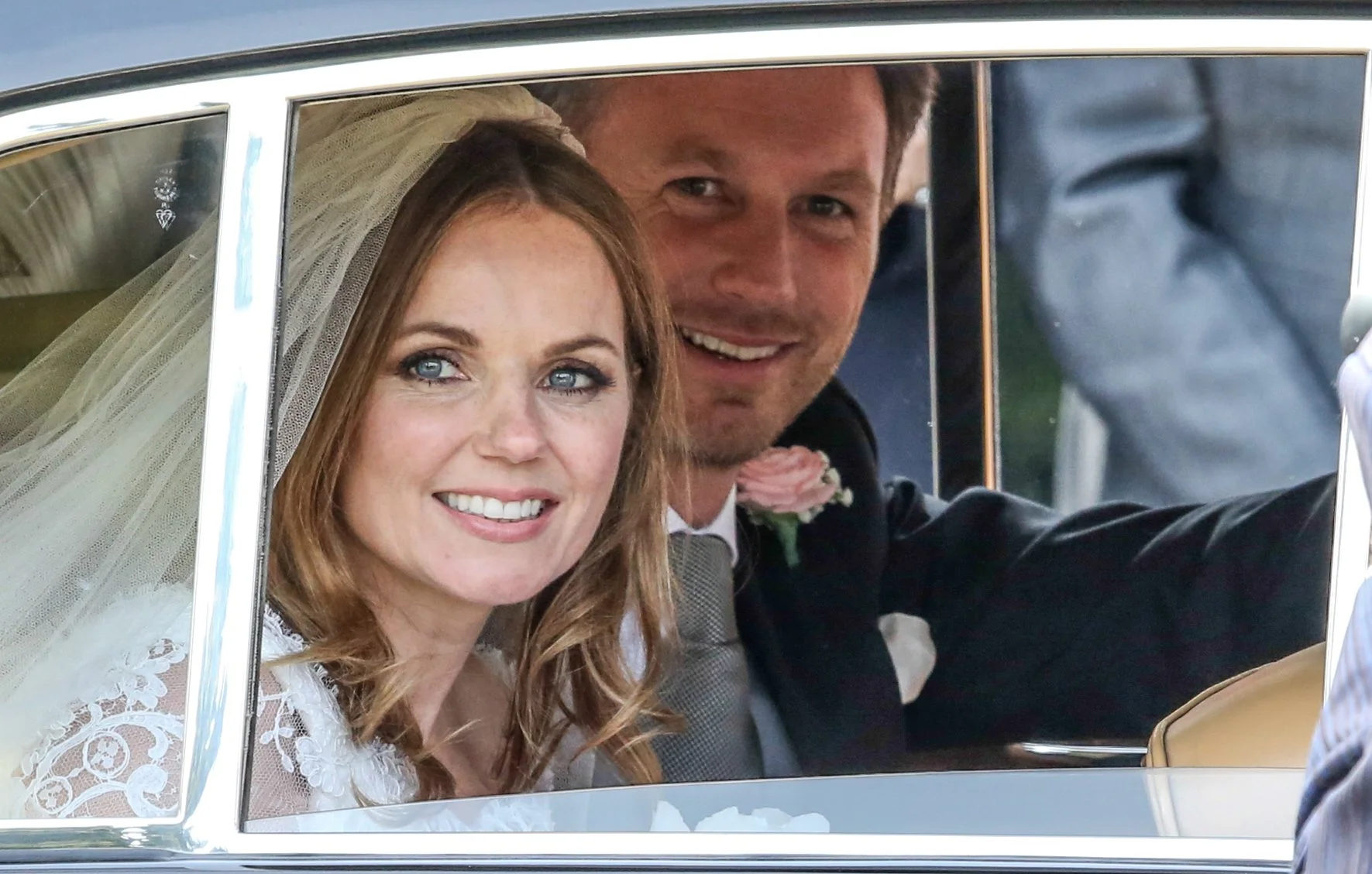 A bride and groom sitting inside a vintage car, smiling and looking out of the window, with the bride wearing a lace veil and the groom in a suit with a pink boutonniere.