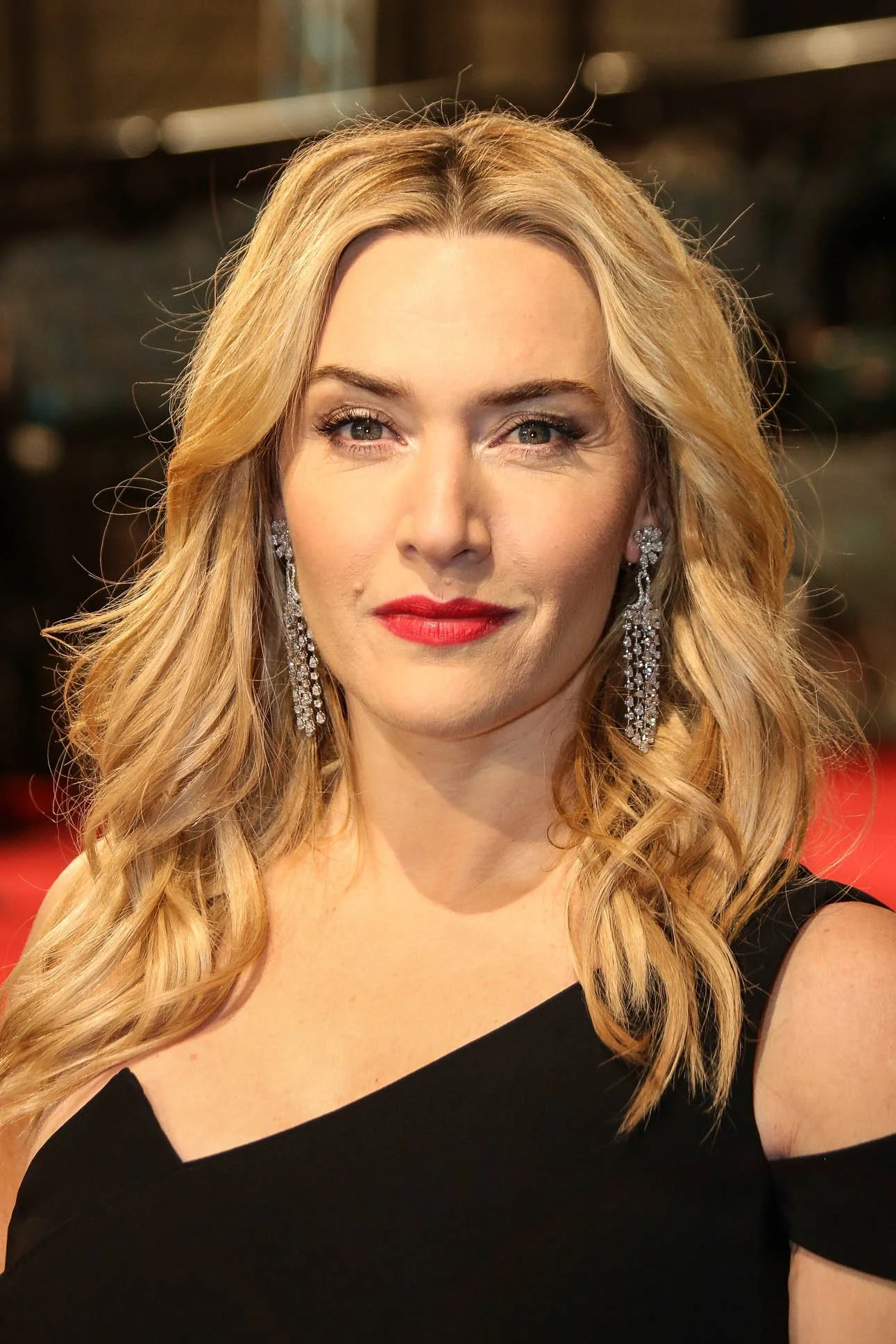 A woman with long, wavy blonde hair wearing a black dress with a one-shoulder design and large, sparkling chandelier earrings.