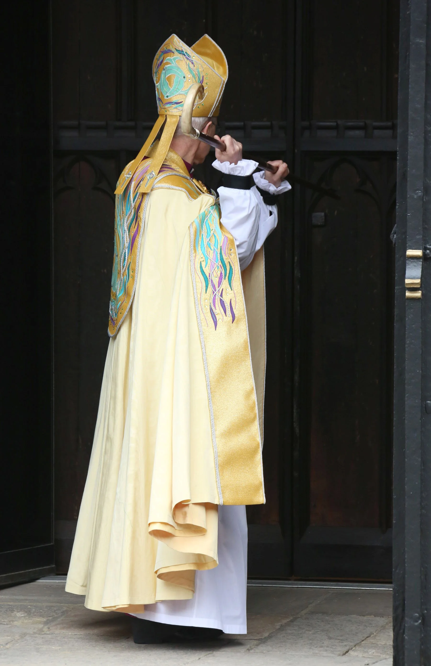 A person dressed in ornate, colorful religious or ceremonial robes holding a staff, standing in front of closed dark wooden doors.