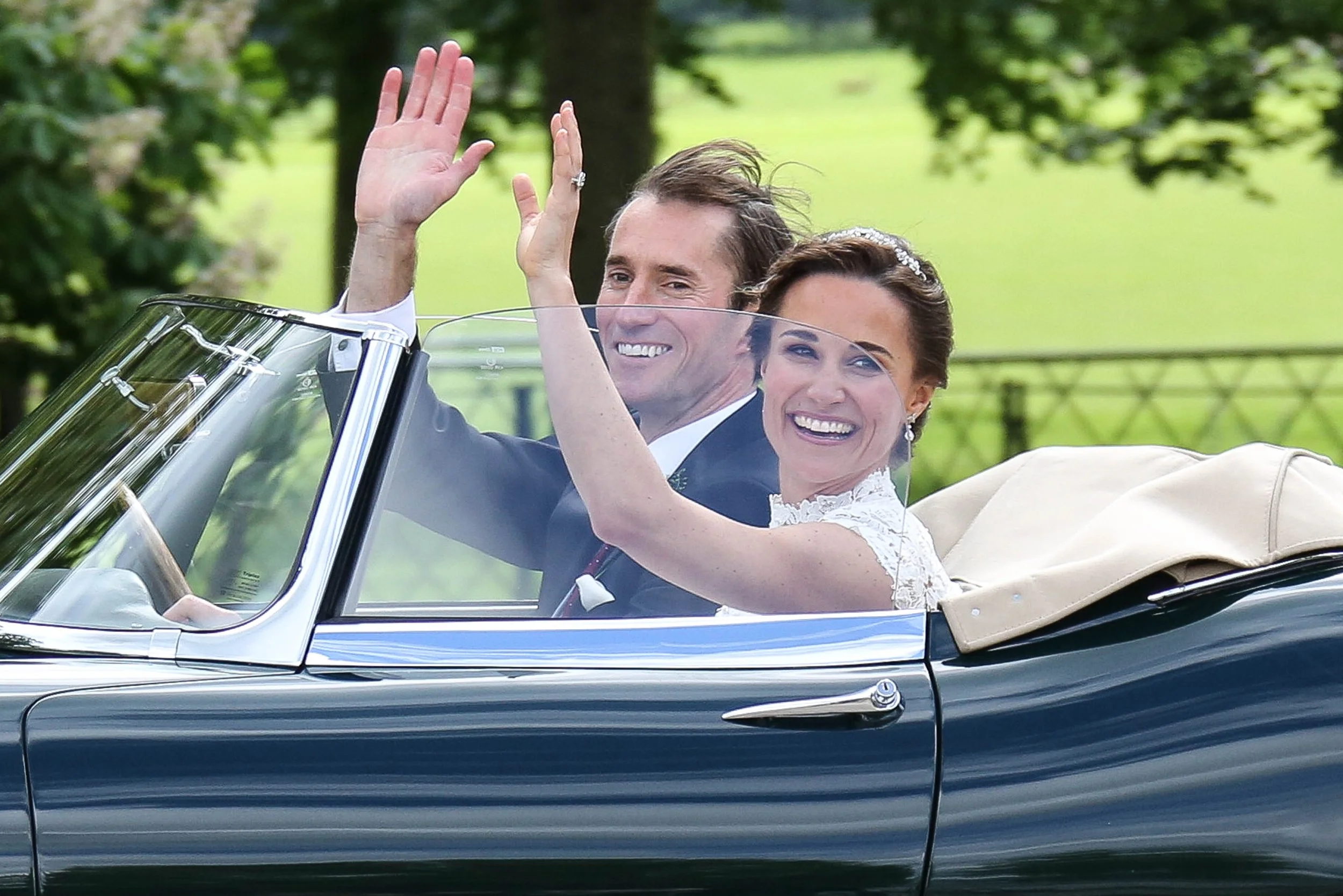 A smiling man and woman in wedding attire riding in a vintage convertible car, waving from the car's windshield, surrounded by a lush green outdoor setting.