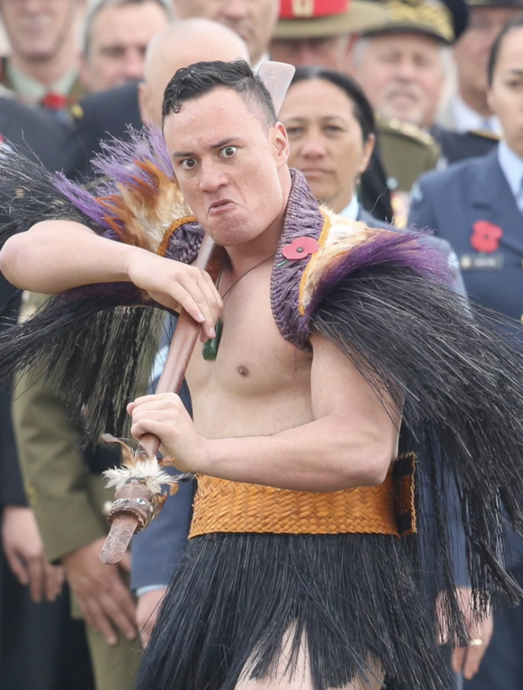 A man in traditional Maori warrior attire holding a weapon with a serious expression, surrounded by people in formal military uniforms at the 100th Anniversary of battle of the Somme NZ Maori Regiment attended by Charles Prince of Wales . France