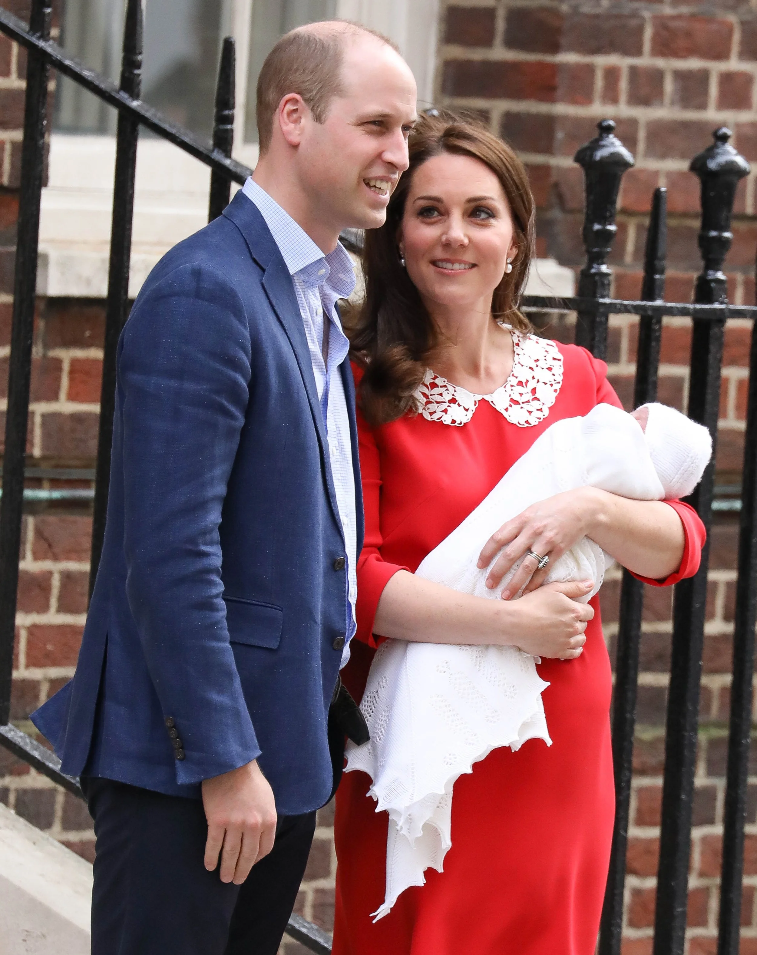 A man in a blue suit and a woman in a red dress holding a baby wrapped in a white blanket, standing outside near a brick building and a black metal fence.