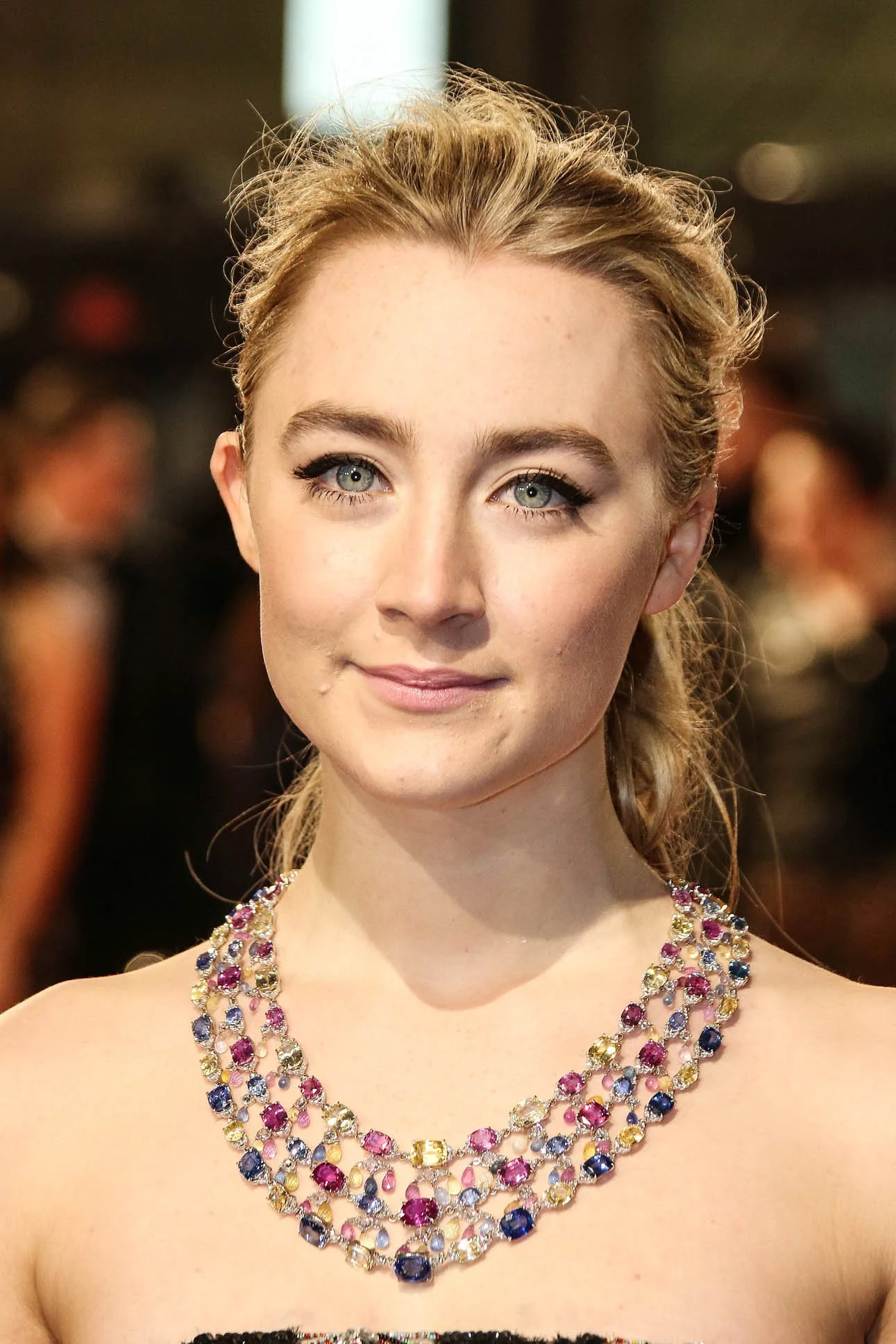 Close-up of a woman with light-colored hair and blue eyes, wearing a colorful multi-layered gemstone necklace, at an indoor event.