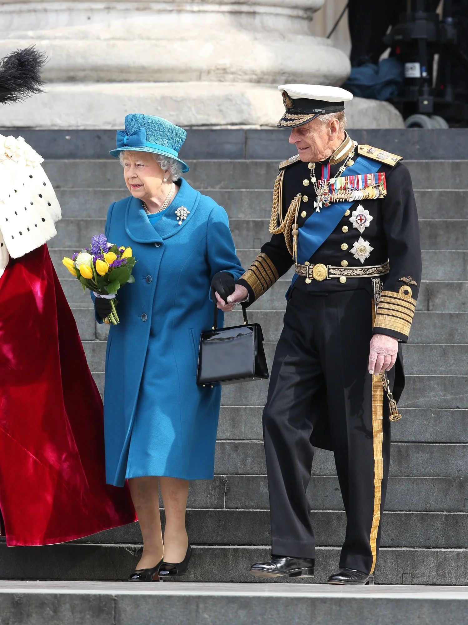 Queen Elizabeth II and Prince Philip holding hands on the steps of a building during a formal event. Queen Elizabeth II is wearing a bright blue coat and matching hat, holding a bouquet of flowers. Prince Philip is dressed in a formal military unifor