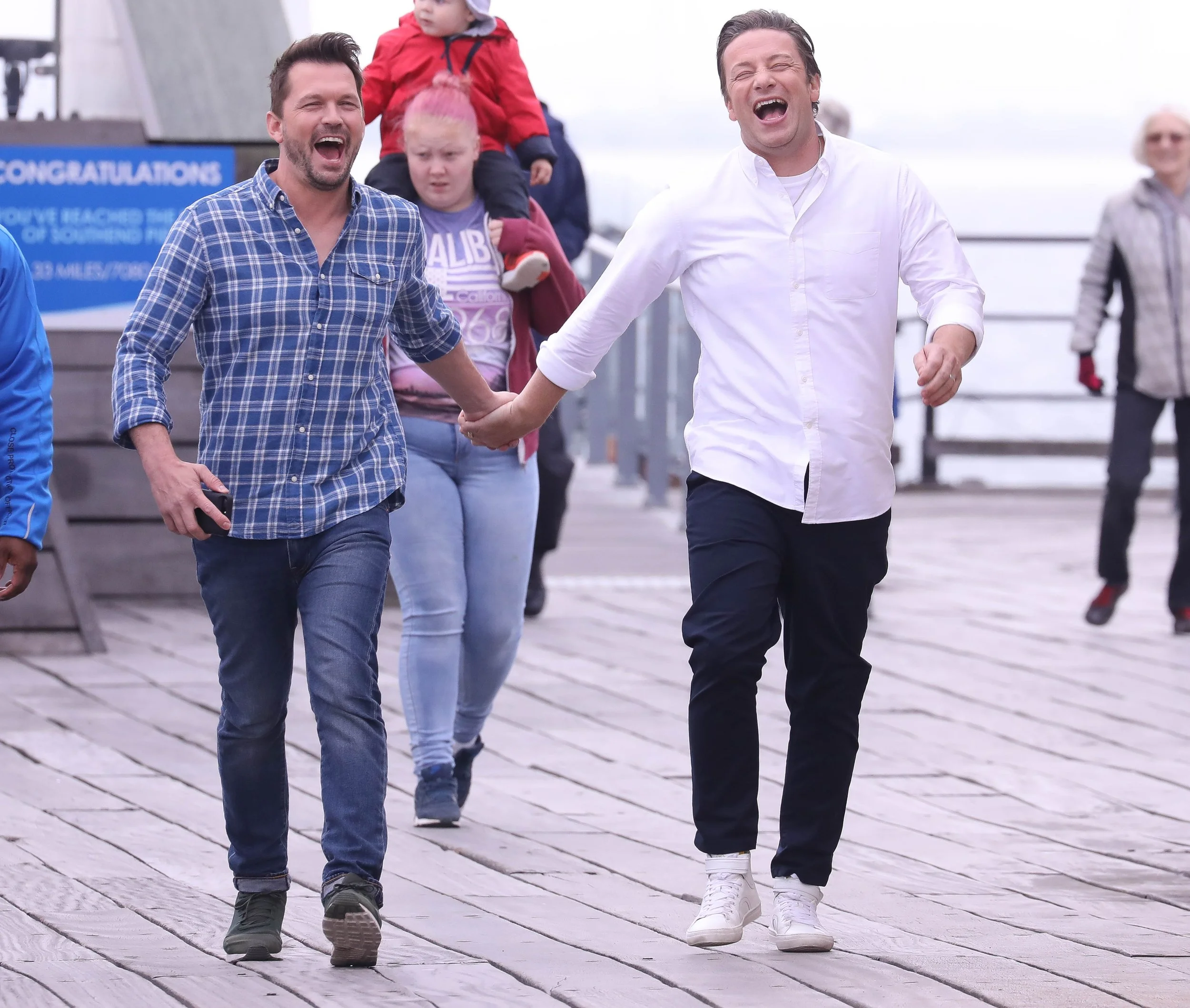 Two men holding hands and laughing while walking on a wooden boardwalk. A young girl with a pink headband and a child in a red jacket are visible in the background.