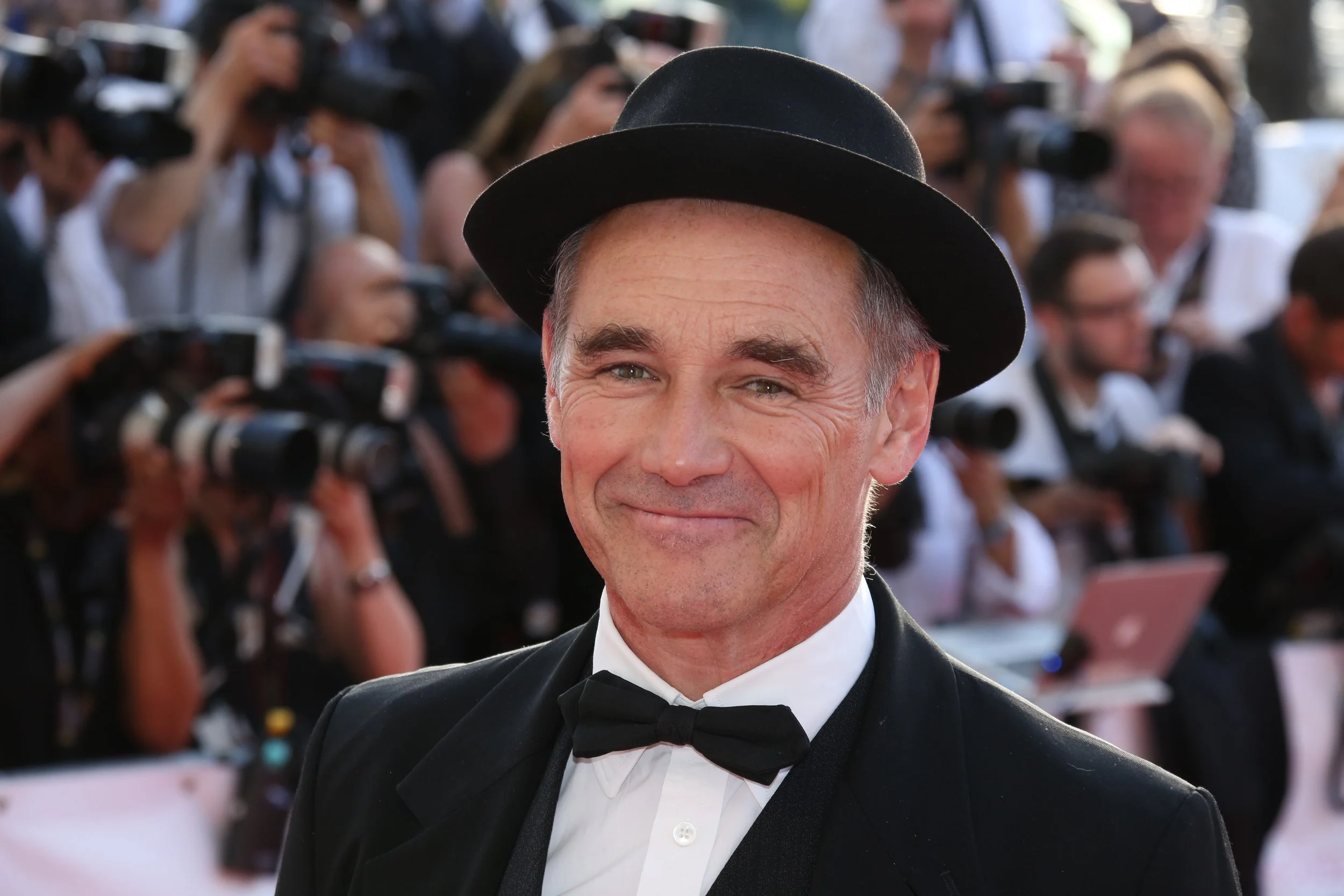 A man in a tuxedo and bow tie smiling on a red carpet, with photographers and people in the background.