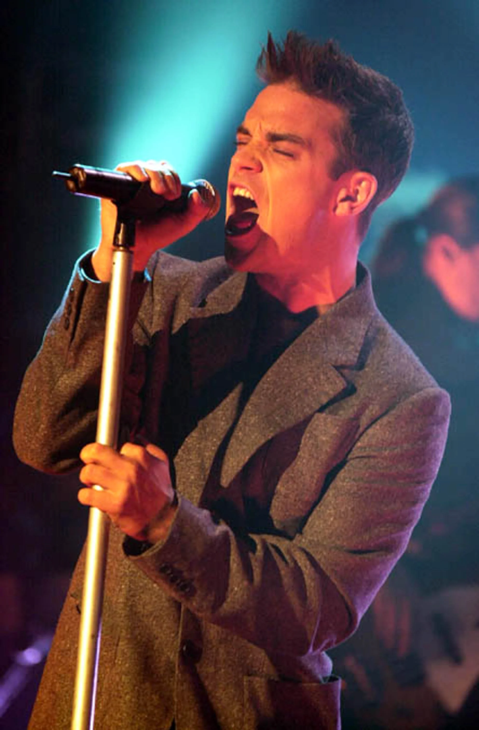 A young man passionately sings into a microphone on stage, wearing a brown blazer and a dark shirt, with a colorful background.